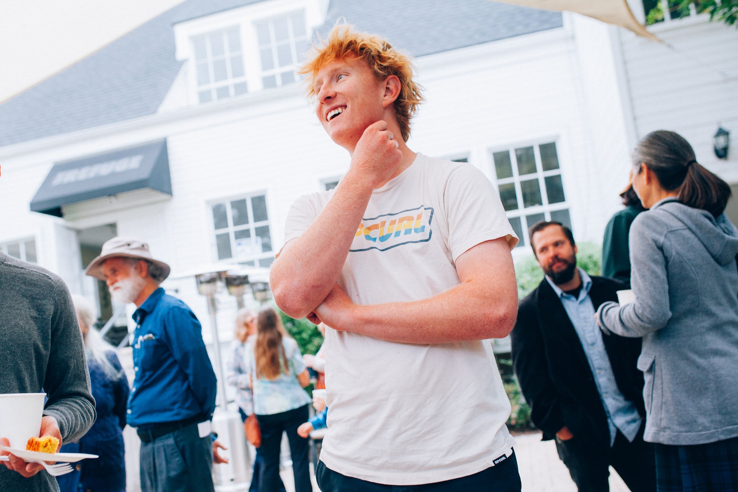 A young man with curly red hair smiles and laughs outdoors at a youth group gathering, with other students and adults nearby