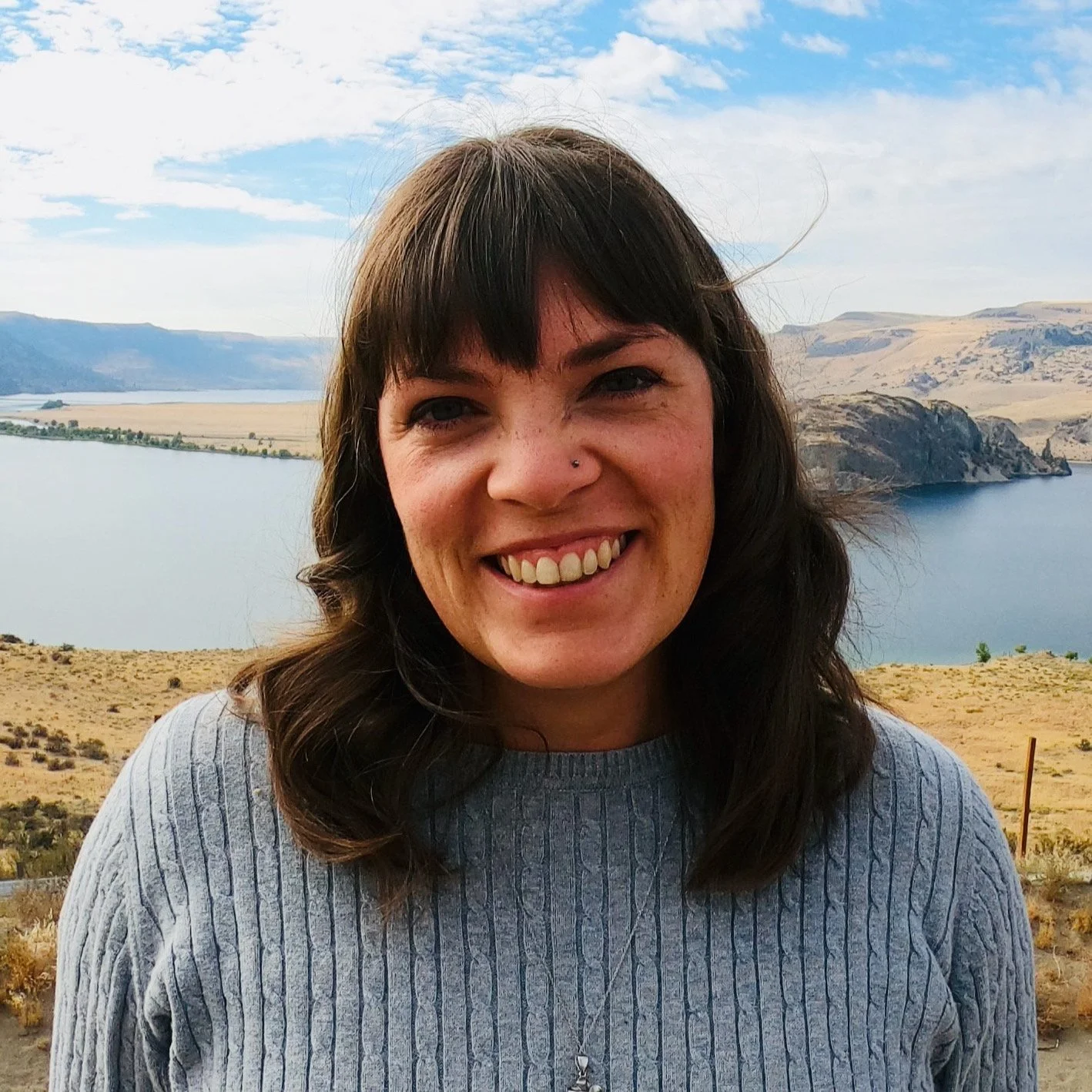 Alayna Dewlen, Ministry Assistant, smiling outdoors with a lake and hills behind her