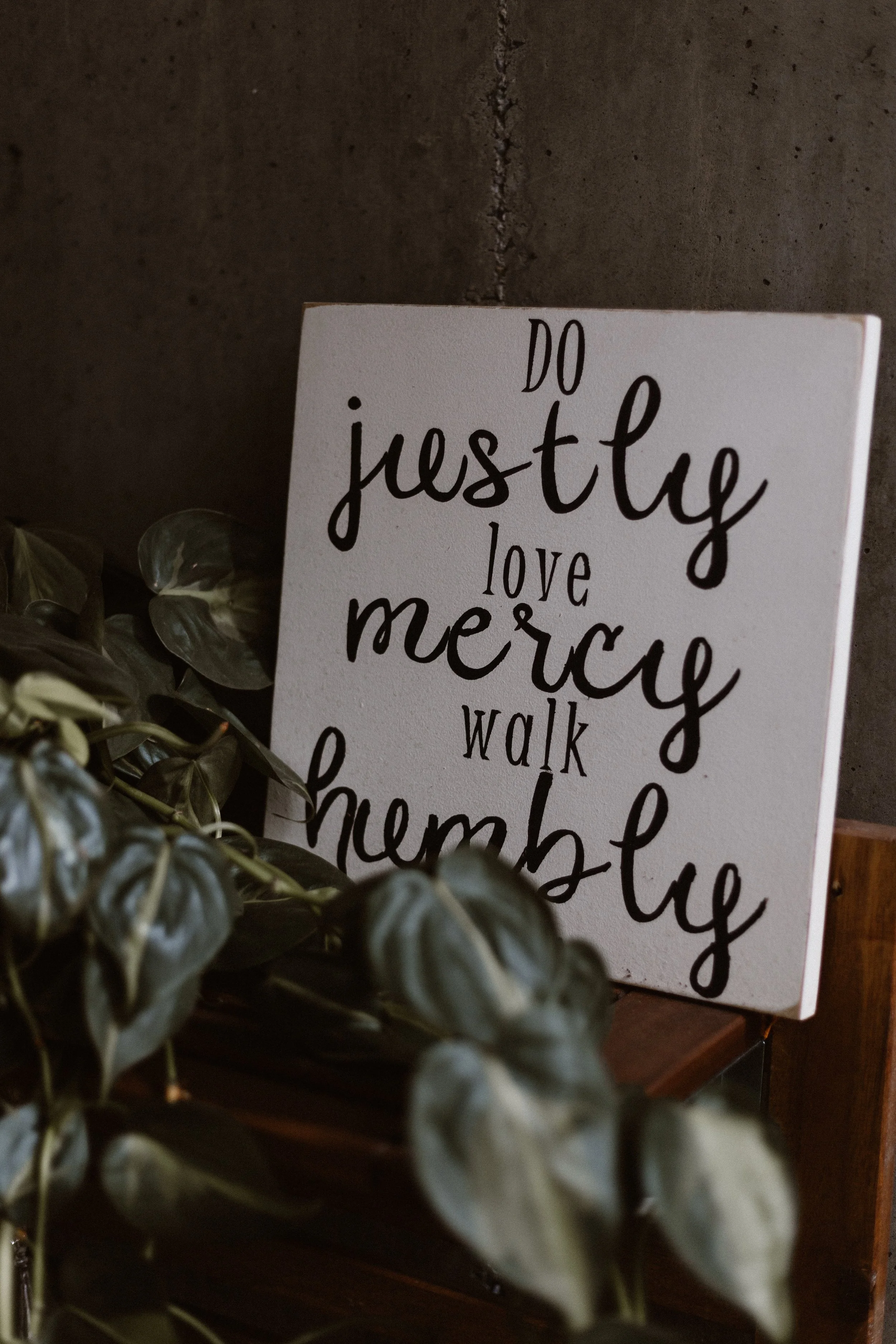 A decorative sign reading "Do justice, love mercy, walk humbly" written in black cursive and serif fonts, placed against a dark textured background, with green leafy plants at the bottom left corner.