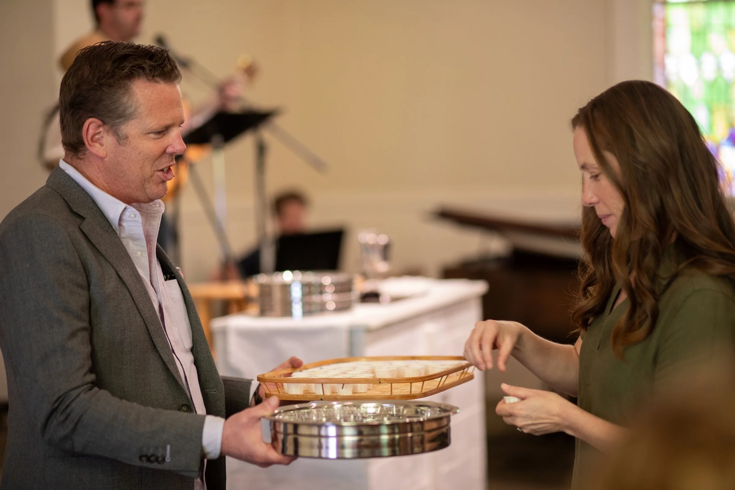 A man in a blazer passes a communion tray to a woman during a worship service, with musicians visible behind them