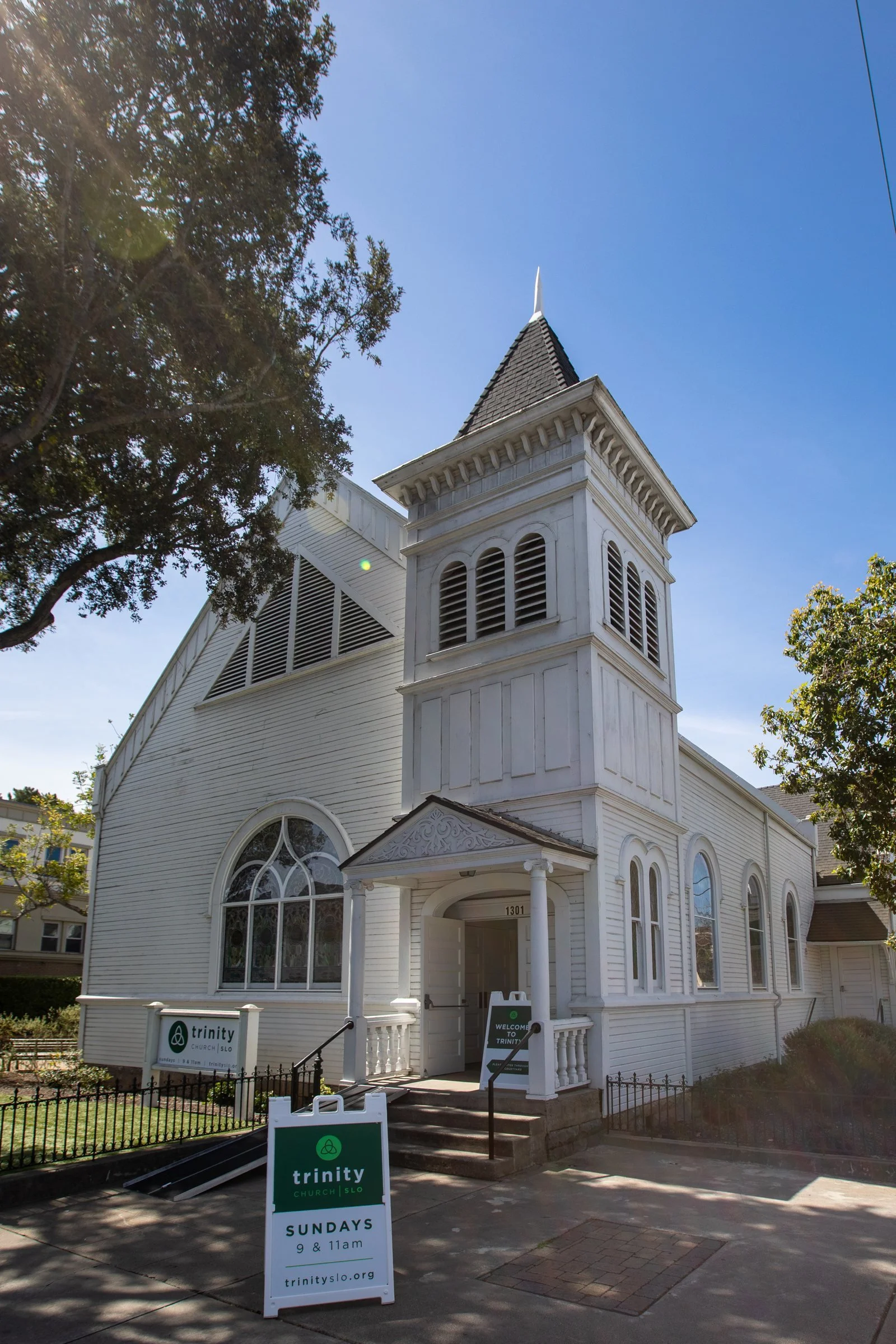 Trinity's white church building with a bell tower, seen from the sidewalk with a Sundays sign out front