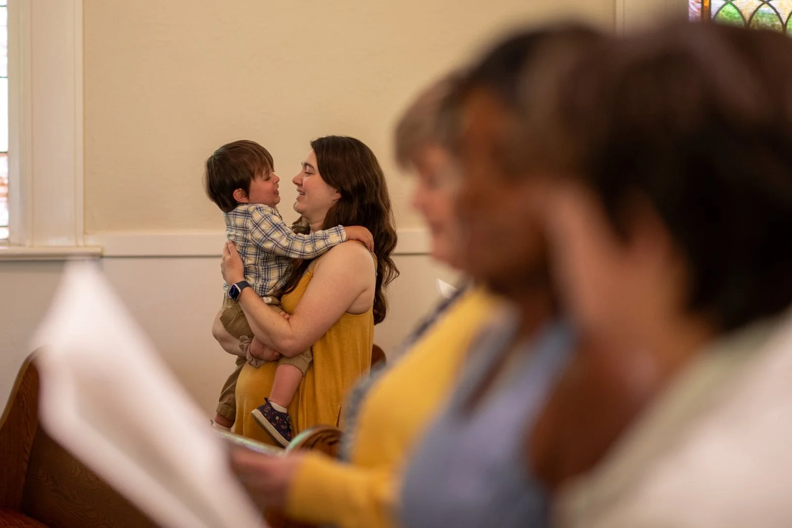 A woman in a yellow top smiles at a toddler in a plaid shirt she is holding, with churchgoers blurred in the foreground