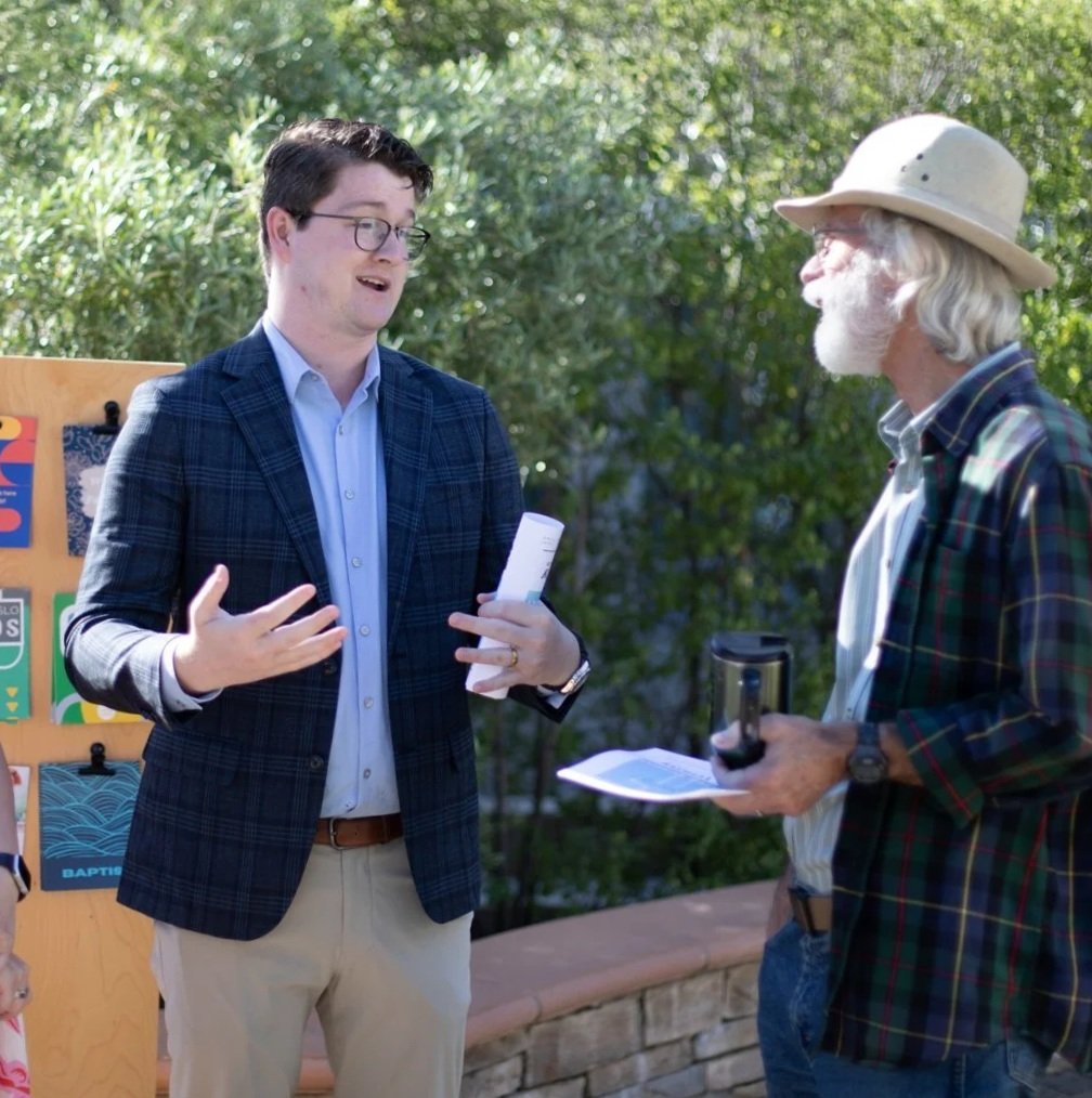 A man in a plaid blazer talks with an older man in a wide-brimmed hat outdoors near a bulletin board