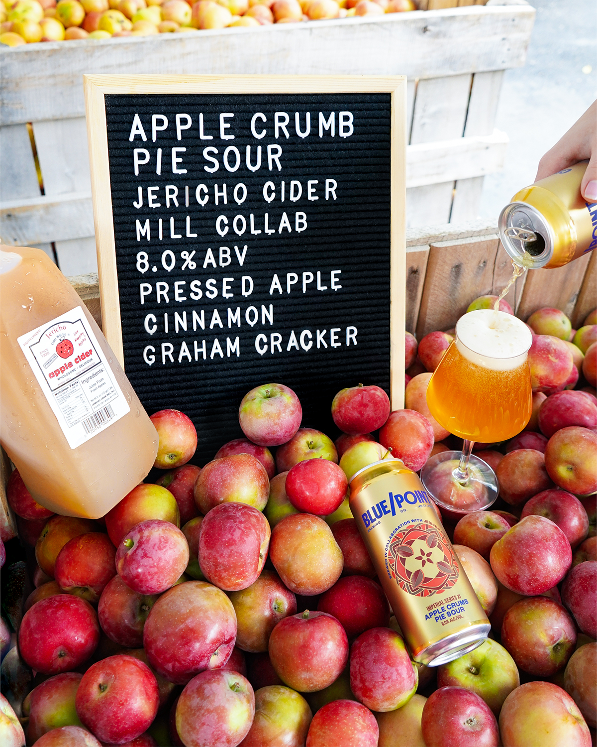 A black letter board displays a list of ingredients for apple crumble pie sour cider, surrounded by red and green apples, with a hand pouring beer into a glass. There are also canned apple cider and a bottle of Blue Point apple sour beer among the ap