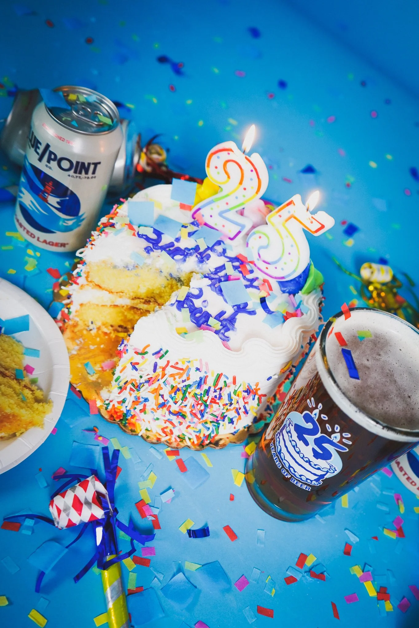 A birthday celebration scene with a slice of cake decorated with rainbow sprinkles, lit candles shaped as the number 25, and surrounded by confetti, party favors, and two cans of beer on a blue tablecloth.