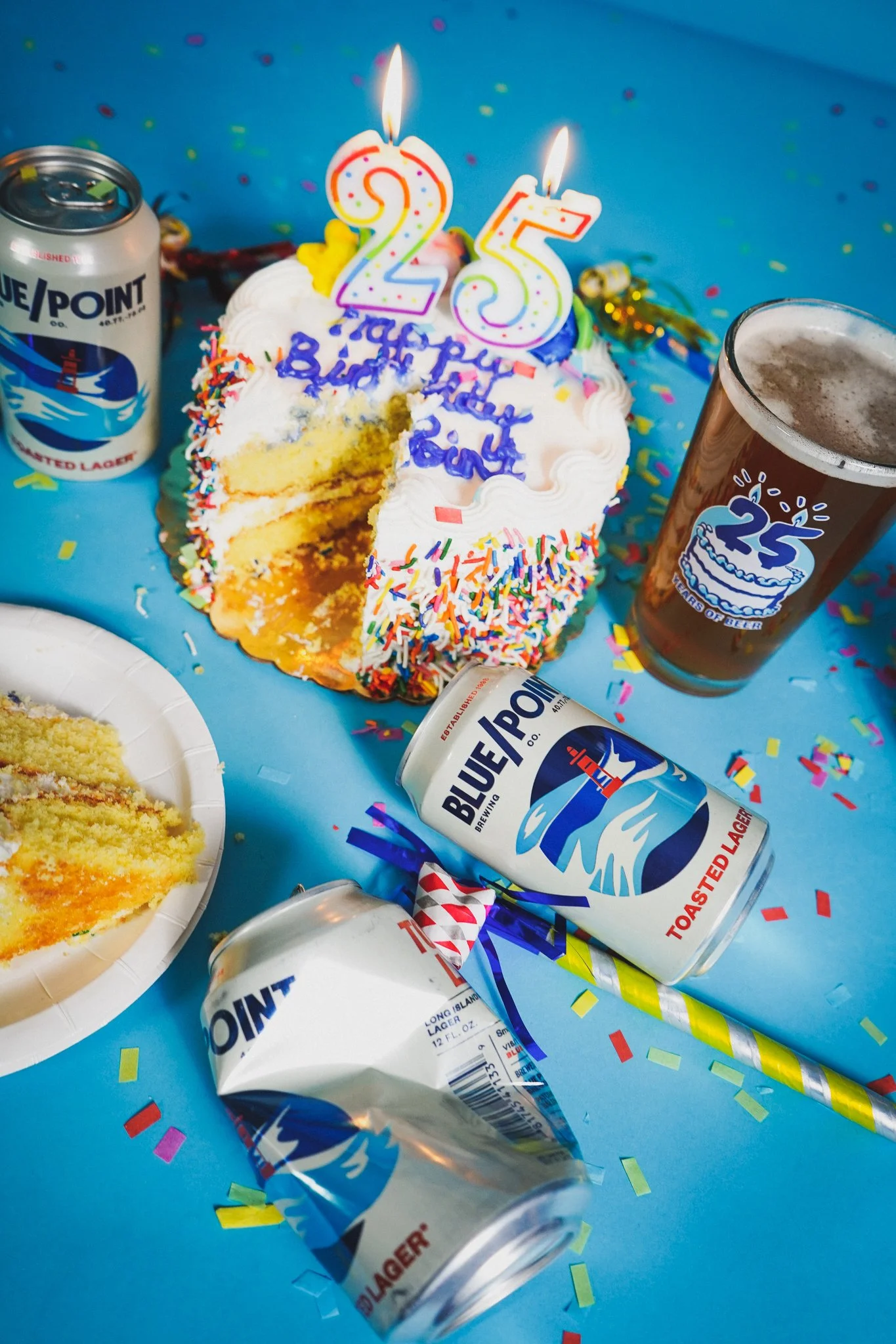 A birthday celebration scene with a cake decorated with colorful sprinkles and two lit candles shaped as the numbers 2 and 5, surrounded by confetti, soda cans, and a glass of beer, on a blue tablecloth.