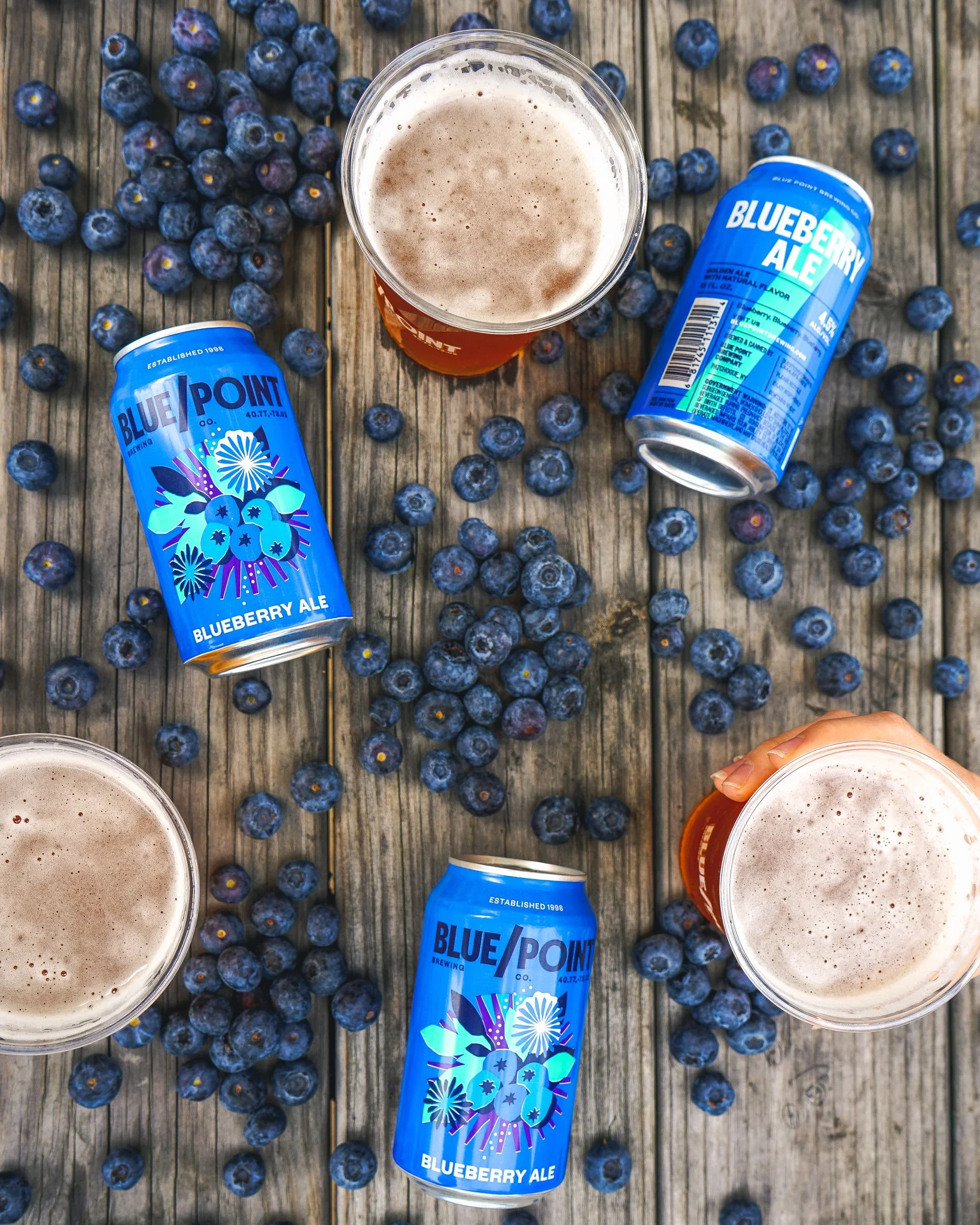 A top-down view of a rustic wooden table with scattered fresh blueberries, two cans of Blueberry Ale from Blue Point Brewing, and two glasses filled with frothy beer.