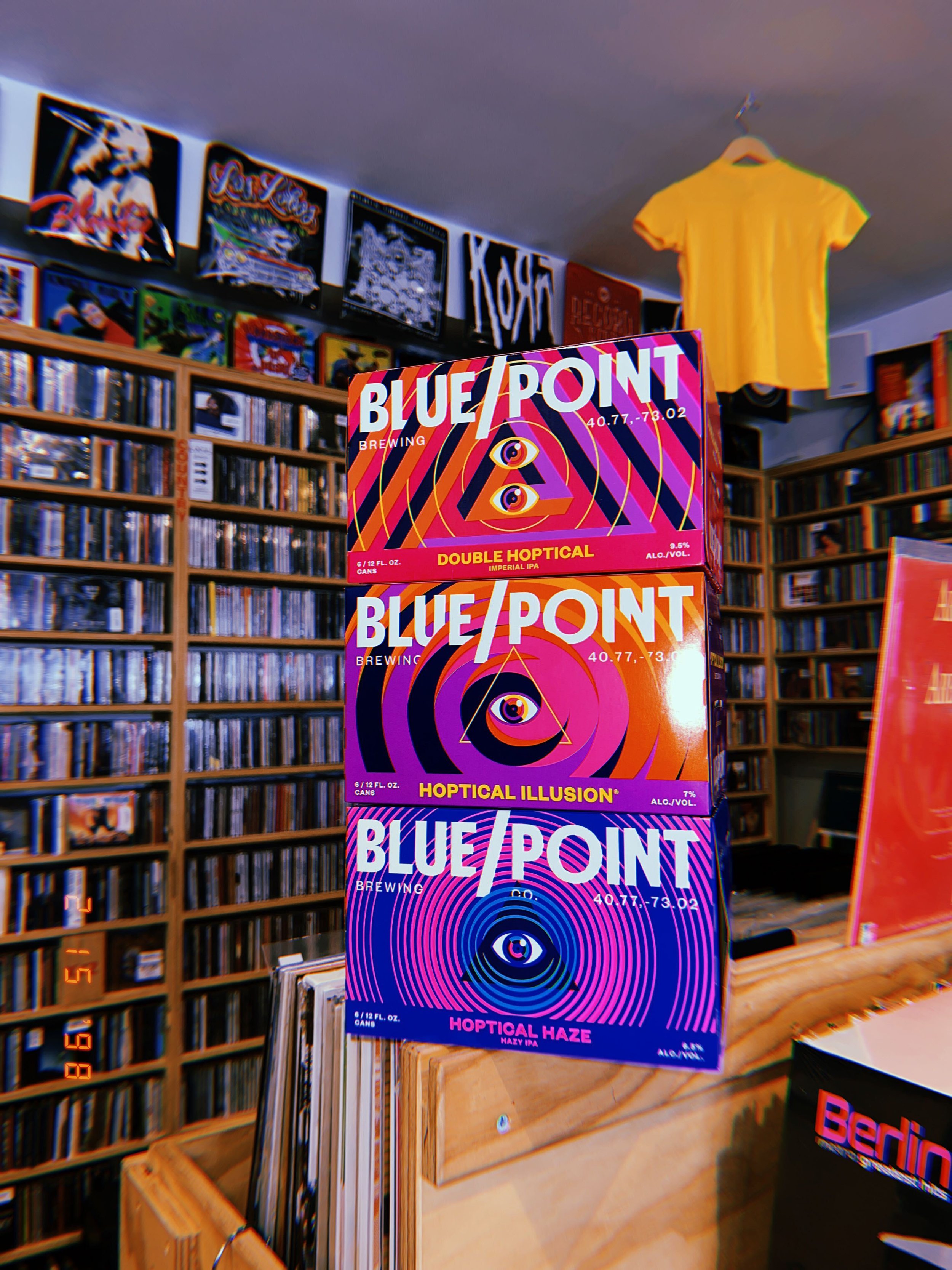 Colorful boxes of Blue Point Brewing Company's Hoptical Illusion and Hazy Haze beers on display in a store, with shelves of DVDs or books in the background.