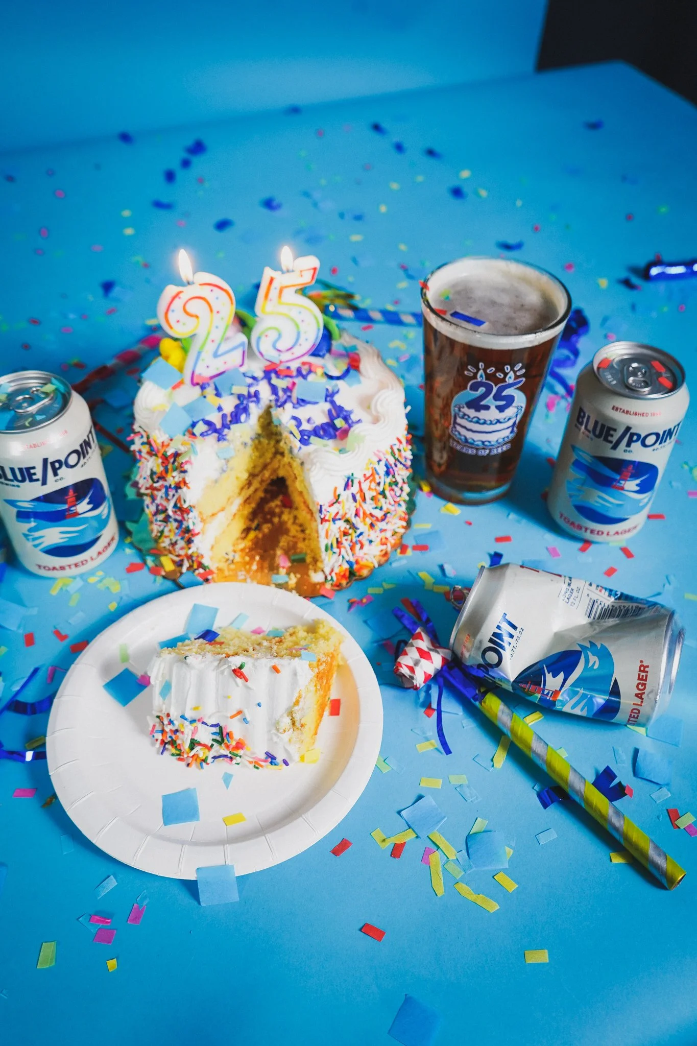 A birthday celebration with a cake decorated with colorful sprinkles and two candles forming the number 25. There are confetti and party supplies, including a plate with a slice of cake, cans of Blue Point beer, and a cup with a 25 logo. The backgrou