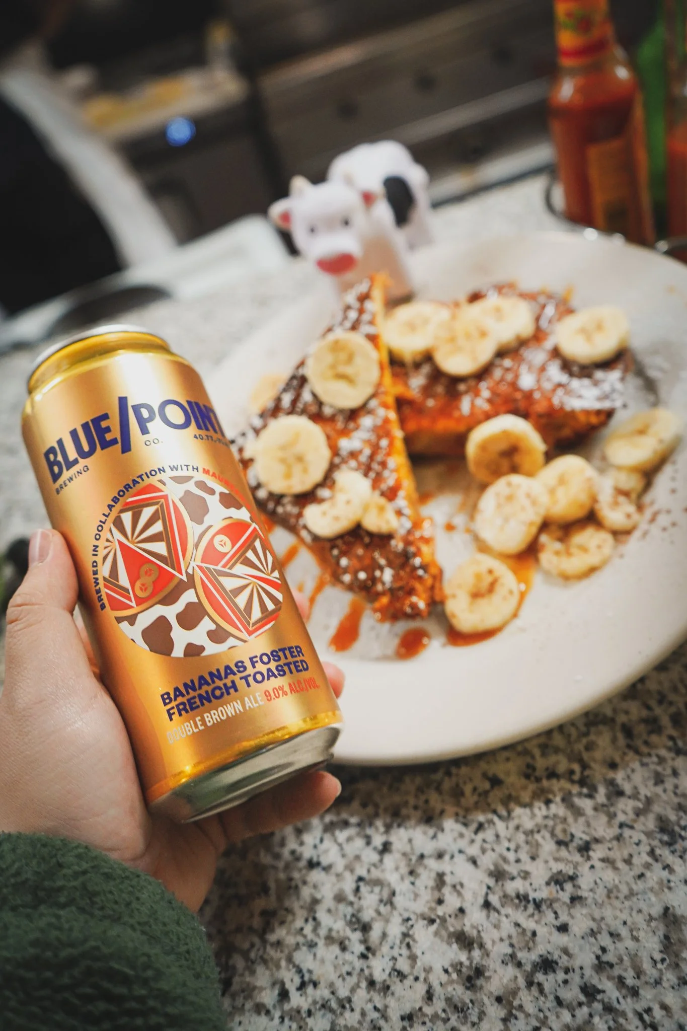 A hand holding a can of Blue Point's bananas foster French toasted double brown ale in front of a plate with French toast topped with banana slices and powdered sugar. The background shows a kitchen setting with bottles and utensils.
