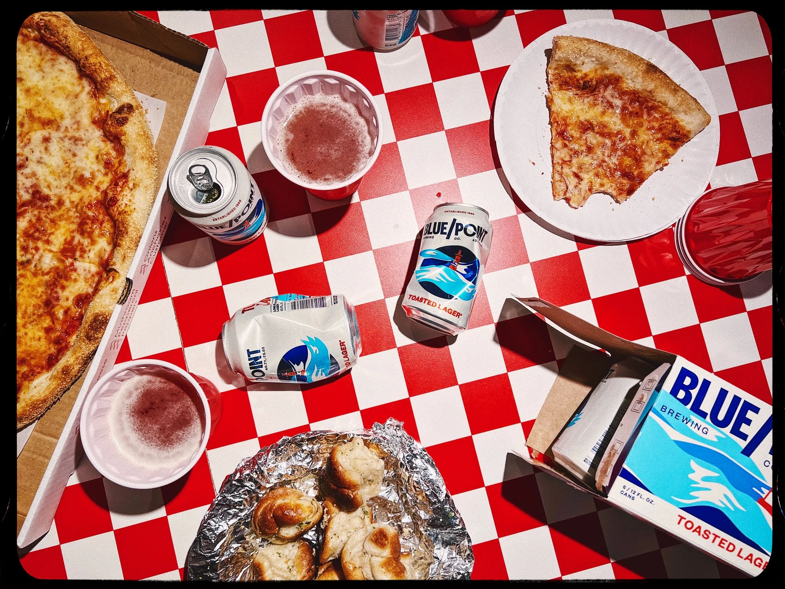 A picnic table with red and white checkered tablecloth, featuring two slices of cheese pizza, a foil-wrapped bread roll, and various beverages including cans and cups of beer and soda, along with a carton of Blue Moon beer.