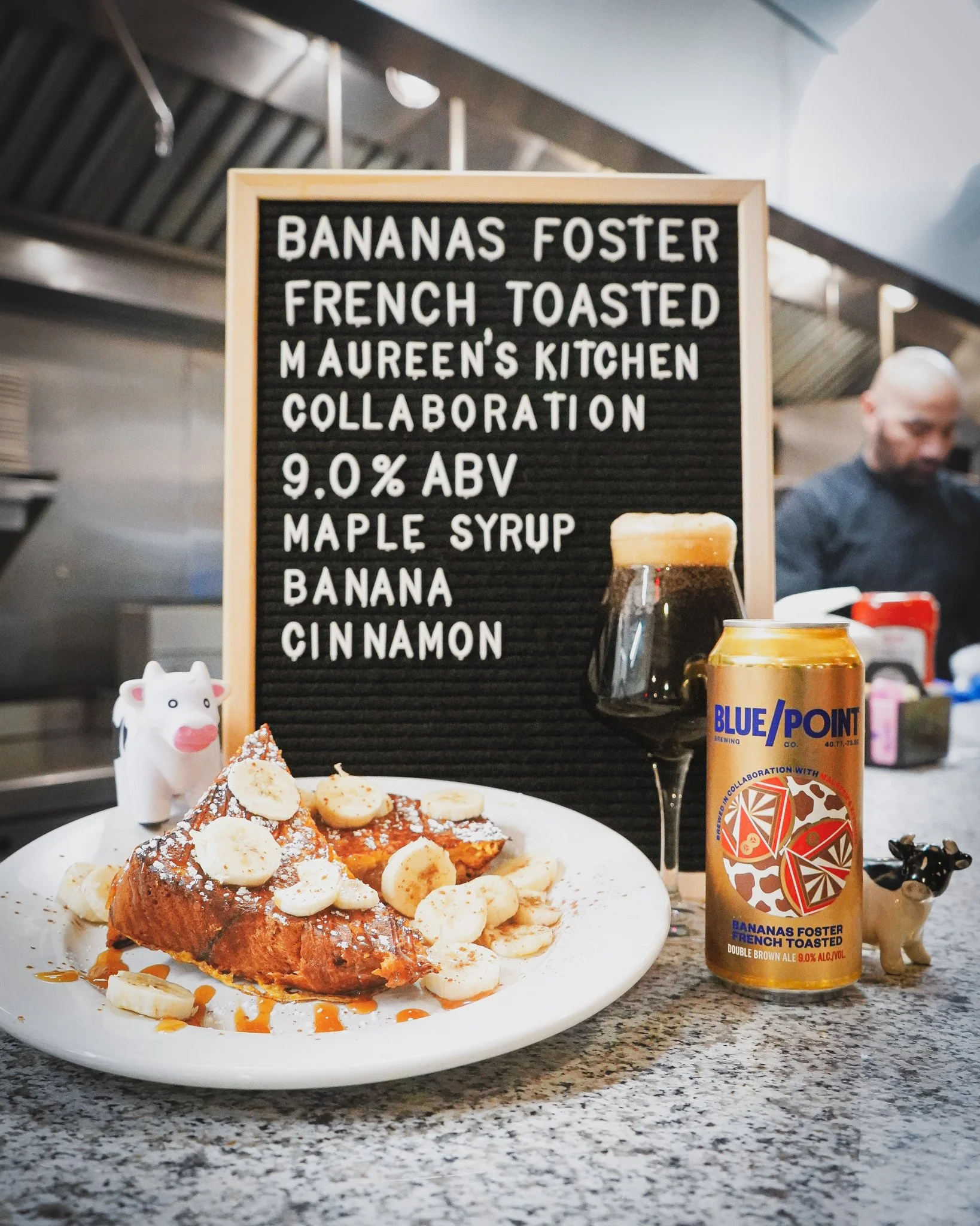 A plate of French toast topped with banana slices, powdered sugar, and syrup in front of a blackboard sign with a list of ingredients and mentions of a collaboration between Bananas Foster and Maureen's Kitchen, and a can of Blue Point beer. In the b