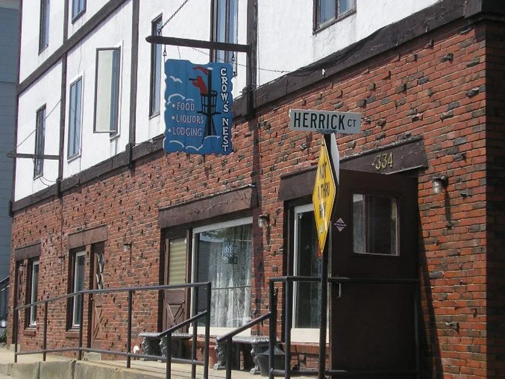 Brick building with signs for a bar, including a sign for Crown's Nest, which sells food, liquor, and lodging, and a street sign for Herrick Court.