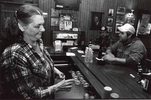 A woman at a bar ordering a drink, with two men sitting at the bar in the background.