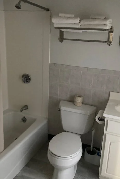 A small bathroom with a bathtub and shower, a toilet with a roll of toilet paper on a stand, a white cabinet, and a wall-mounted towel rack with neatly folded towels.