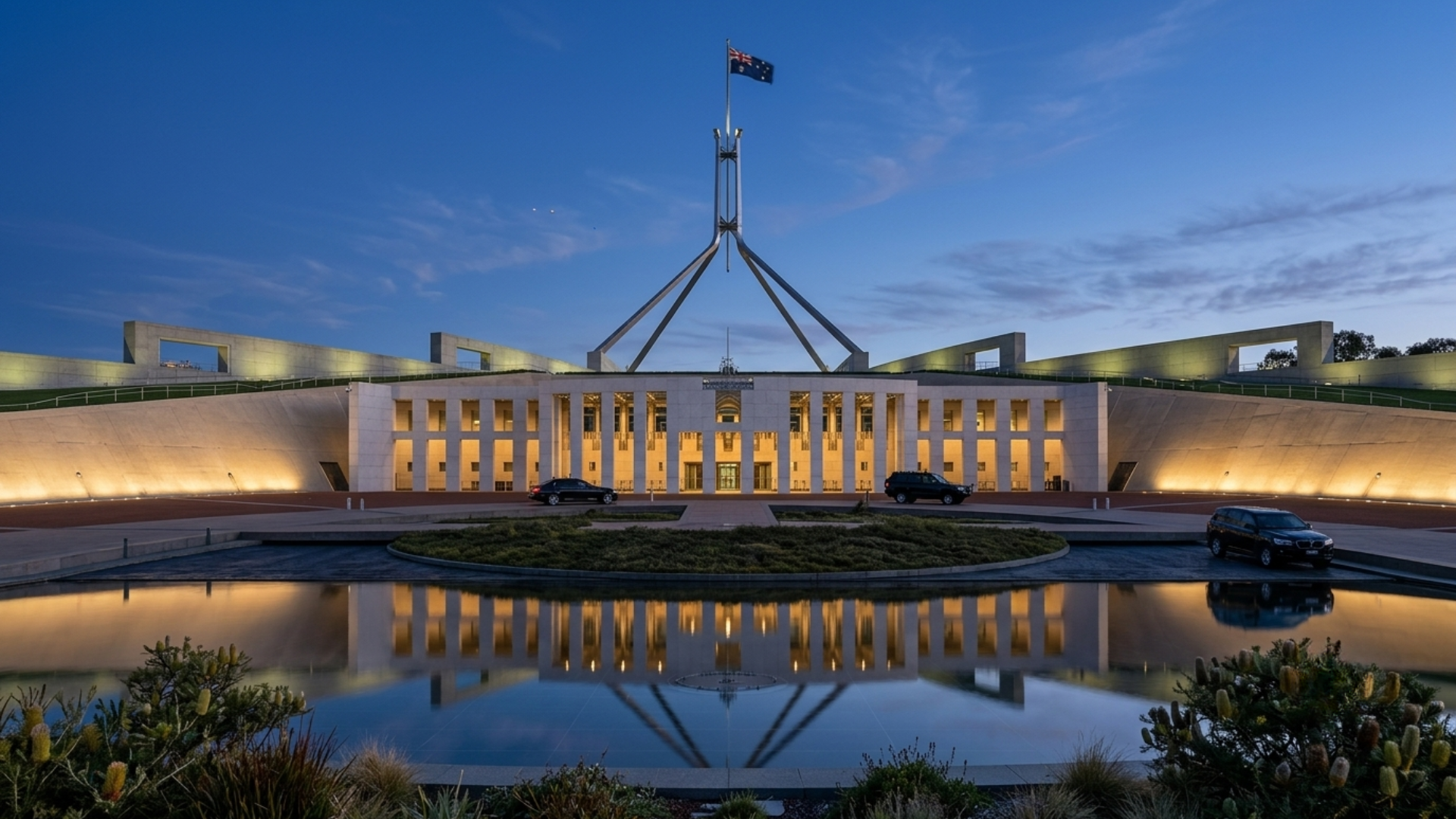 The Australian Parliament House at dusk with illuminated interior lights and a reflection in a water feature in front.