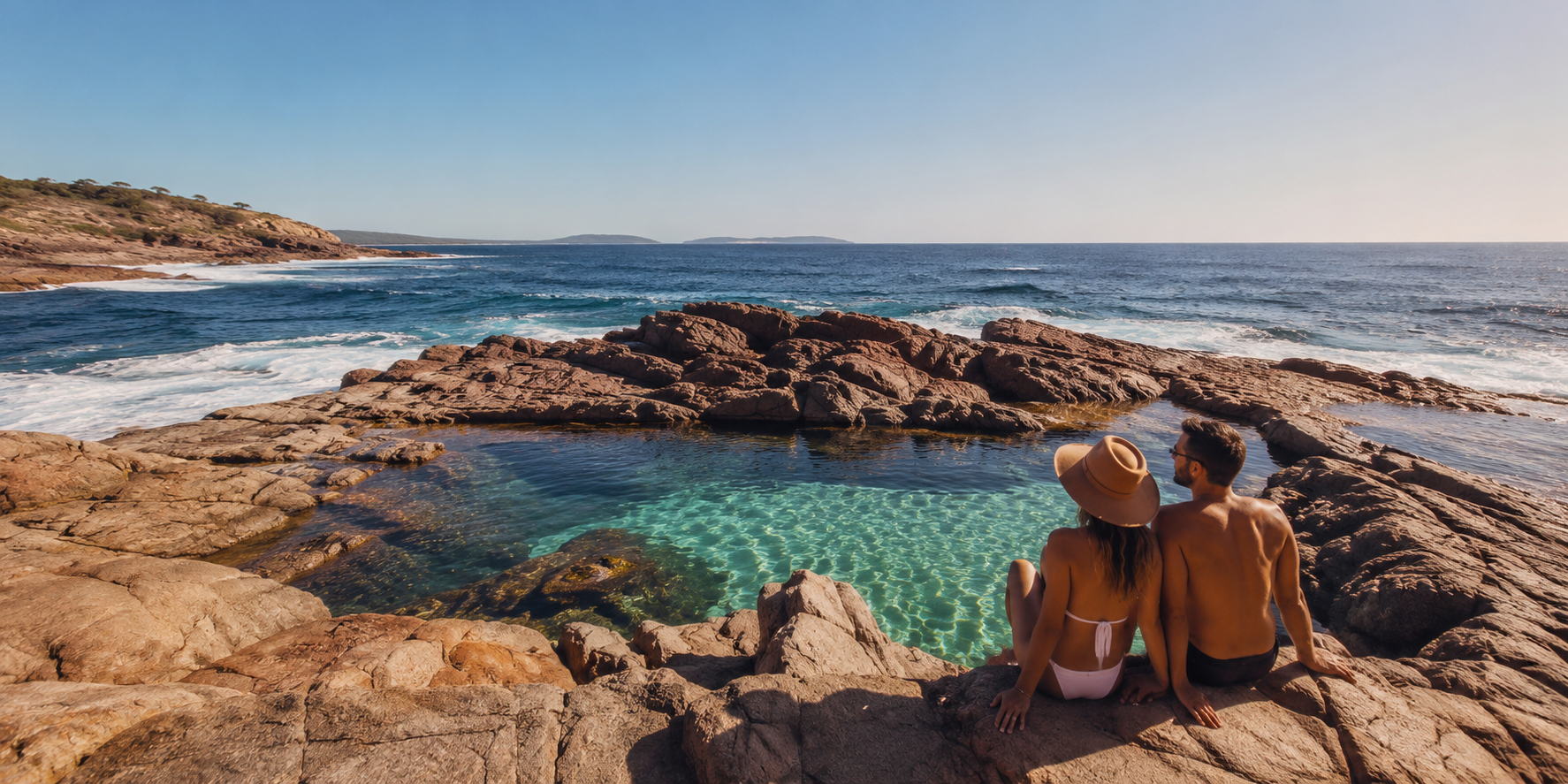A couple sitting on rocks by a natural ocean pool, with the ocean and a distant shoreline in the background on a sunny day.