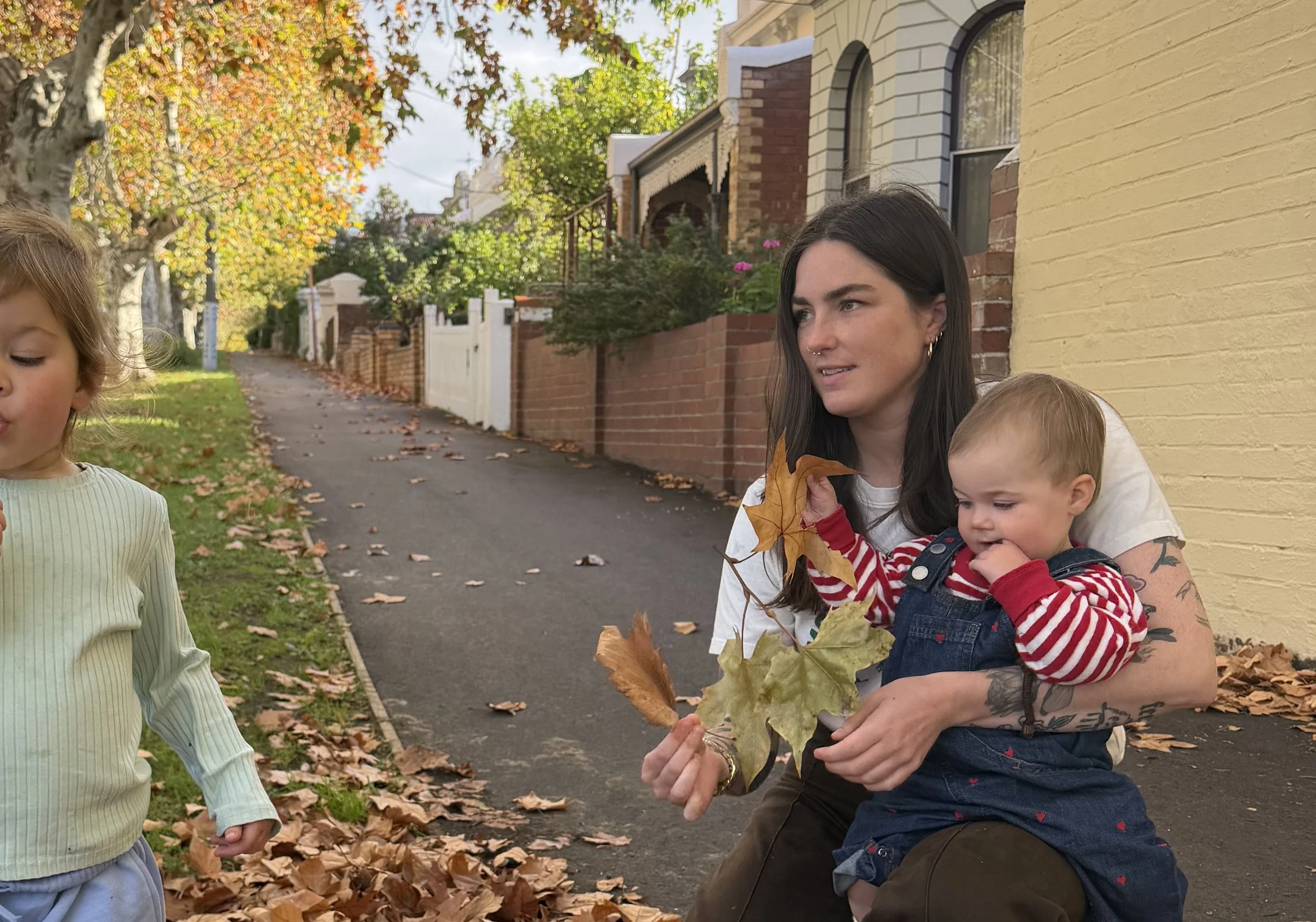 Carli outside in autumn hodling leaves and a baby wearing a red striped long sleeve and demin overalls, looking at a small child with green long sleeve and jeans