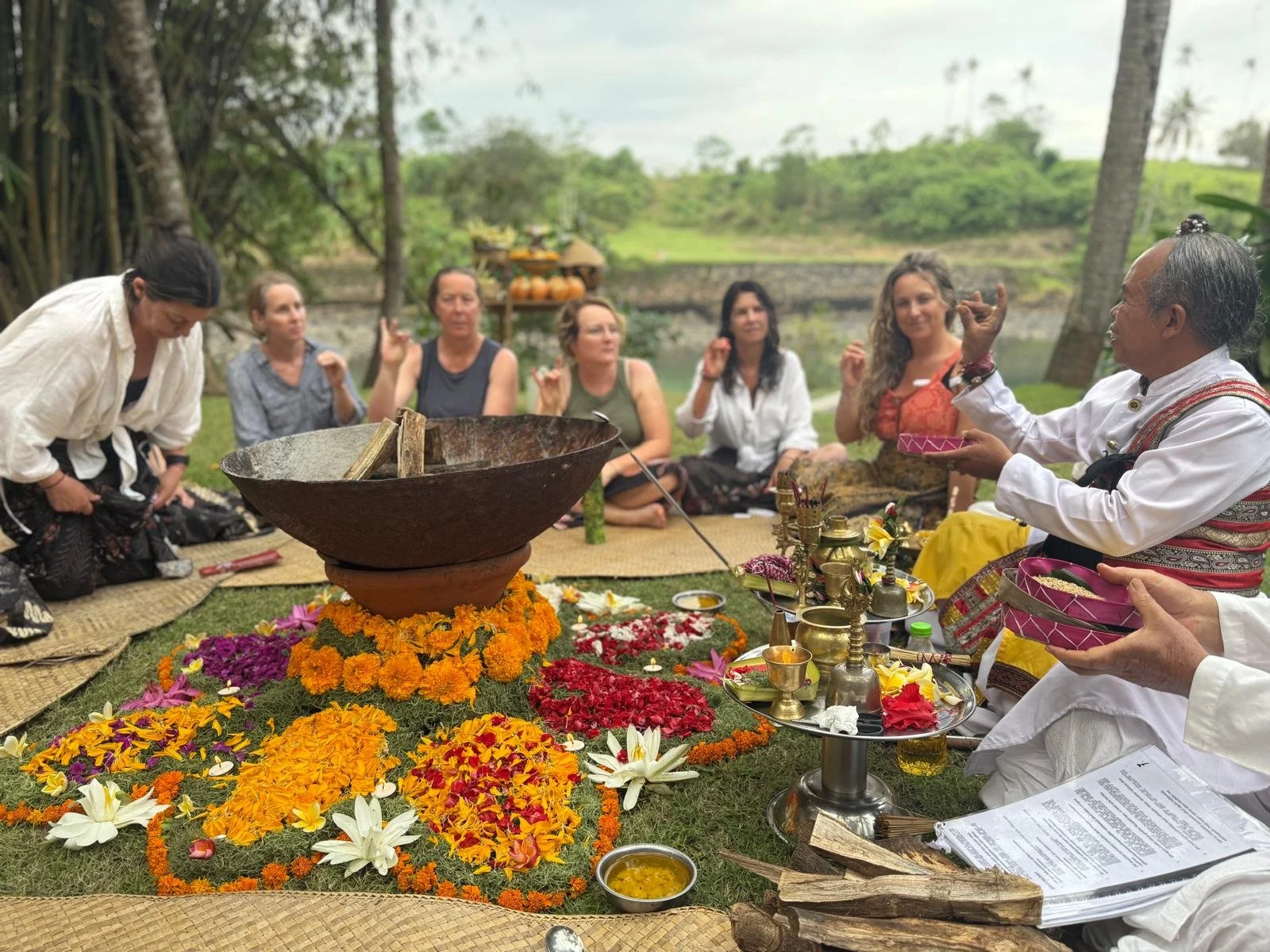 People participating in a traditional outdoor ceremony or ritual, sitting on mats and surrounded by flowers, with a man in traditional attire speaking and holding bowls, in a lush natural setting.