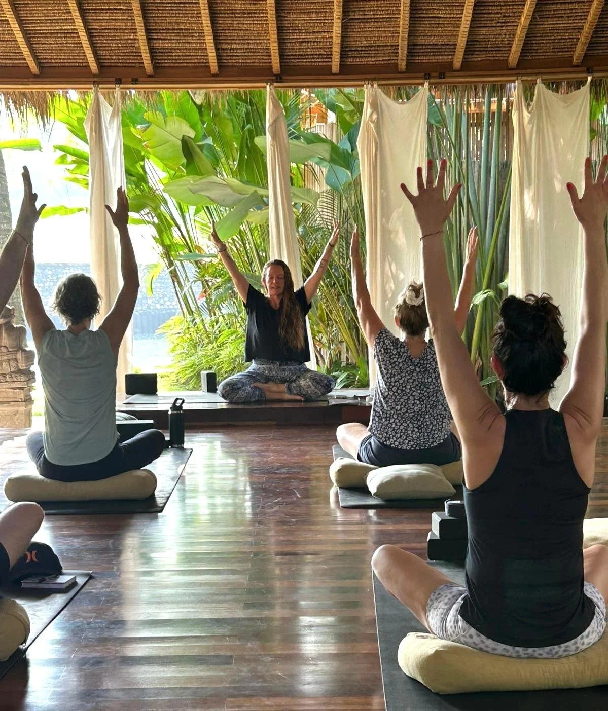 People participating in a yoga or meditation class indoors, with a woman leading, seated cross-legged on a platform, surrounded by lush greenery and large windows.