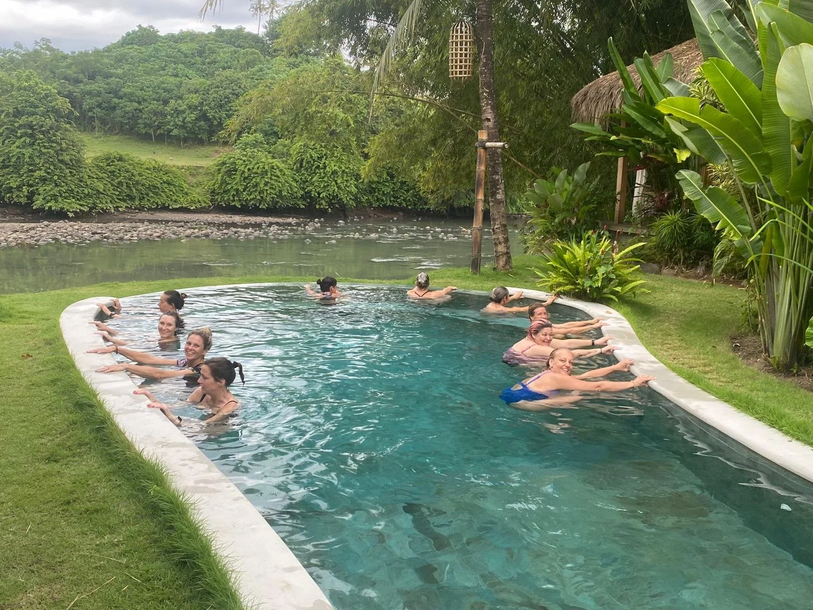 People enjoying a group yoga or exercise session in a small pool or hot tub outdoors surrounded by lush green tropical plants and trees.