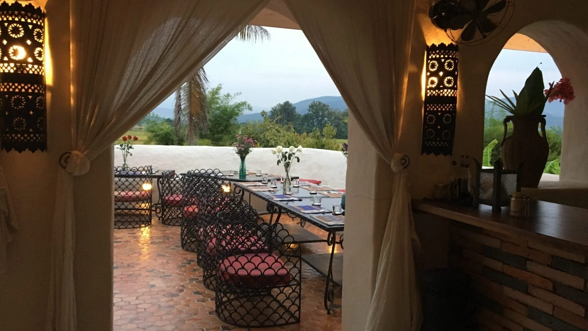Interior of a restaurant with a long dining table, decorated with flowers, set for a meal, overlooking a lush green landscape and mountains outside through an open archway.