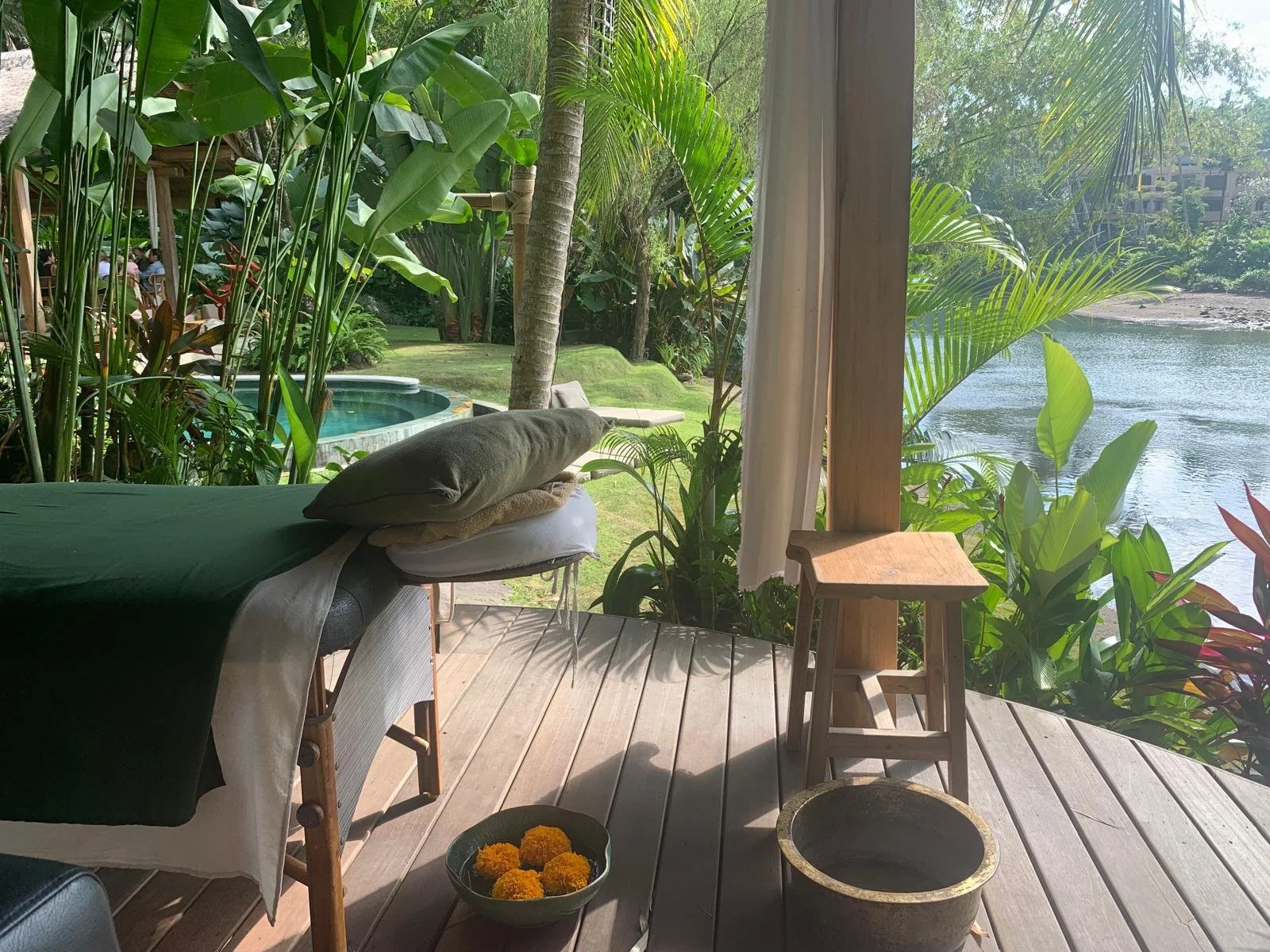 A tranquil outdoor spa setup on a wooden deck overlooking a river, with lush green tropical plants, a massage table with cushions, and a view of a pool and garden area beyond.