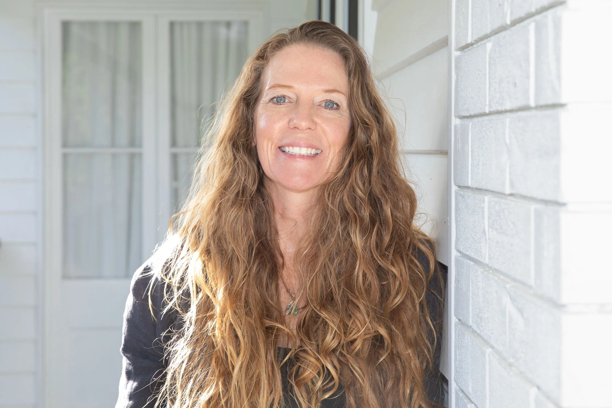 A woman with long, curly, light brown hair smiling and standing next to a white brick wall outside a house with a window in the background.