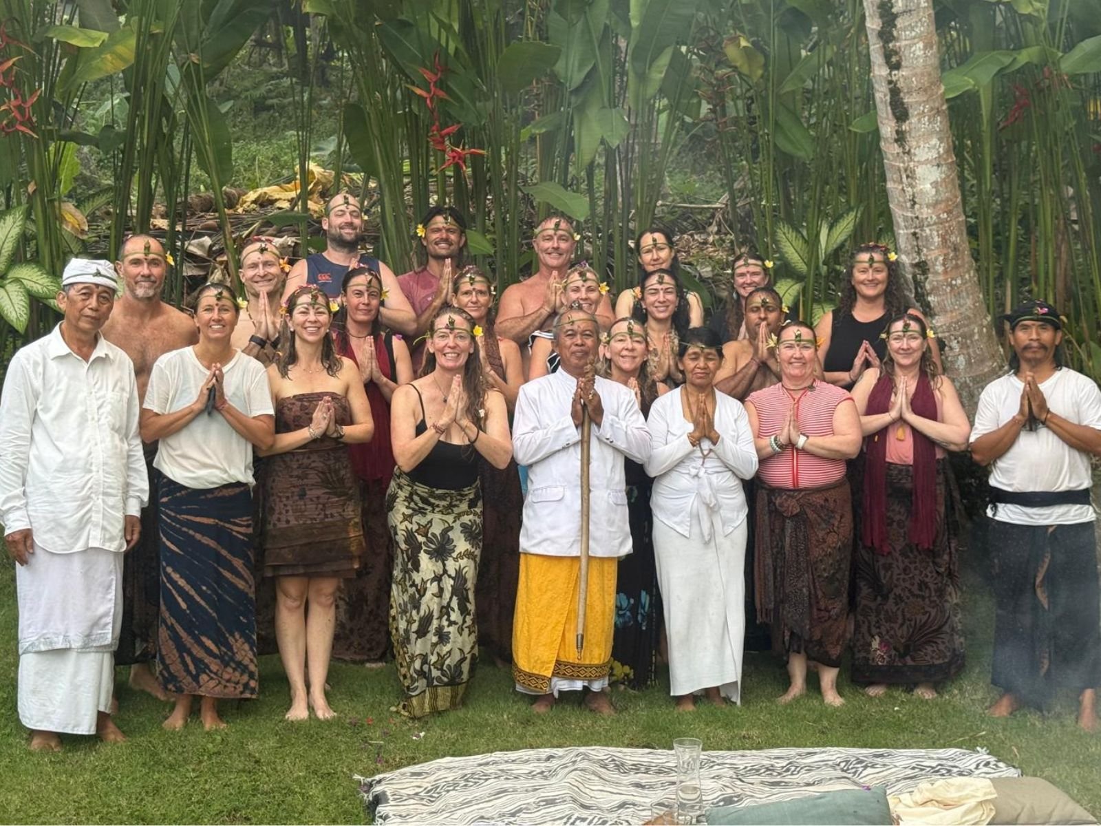Group of people in traditional attire standing outdoors in front of lush green foliage, some with hands in prayer position, others smiling, with a mat on the ground in front.