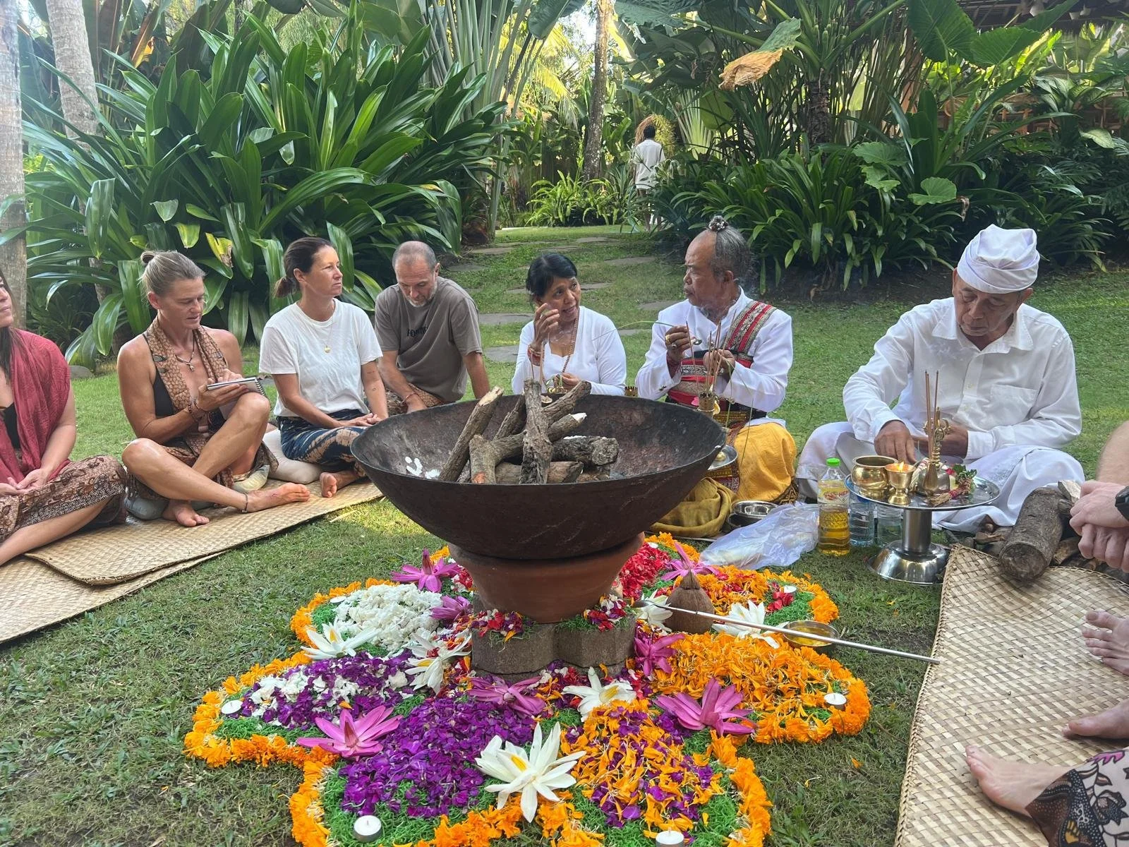 Group of people participating in a traditional outdoor ceremony, sitting on the grass around a large flame with colorful flower decorations, surrounded by lush green plants and trees.