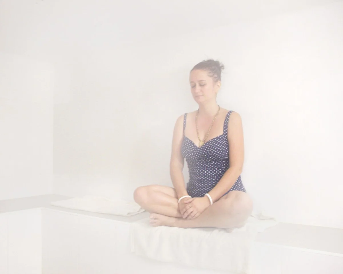 A woman sitting cross-legged with eyes closed against a plain white wall, dressed in a navy polka dot top, wearing necklaces and bracelets.