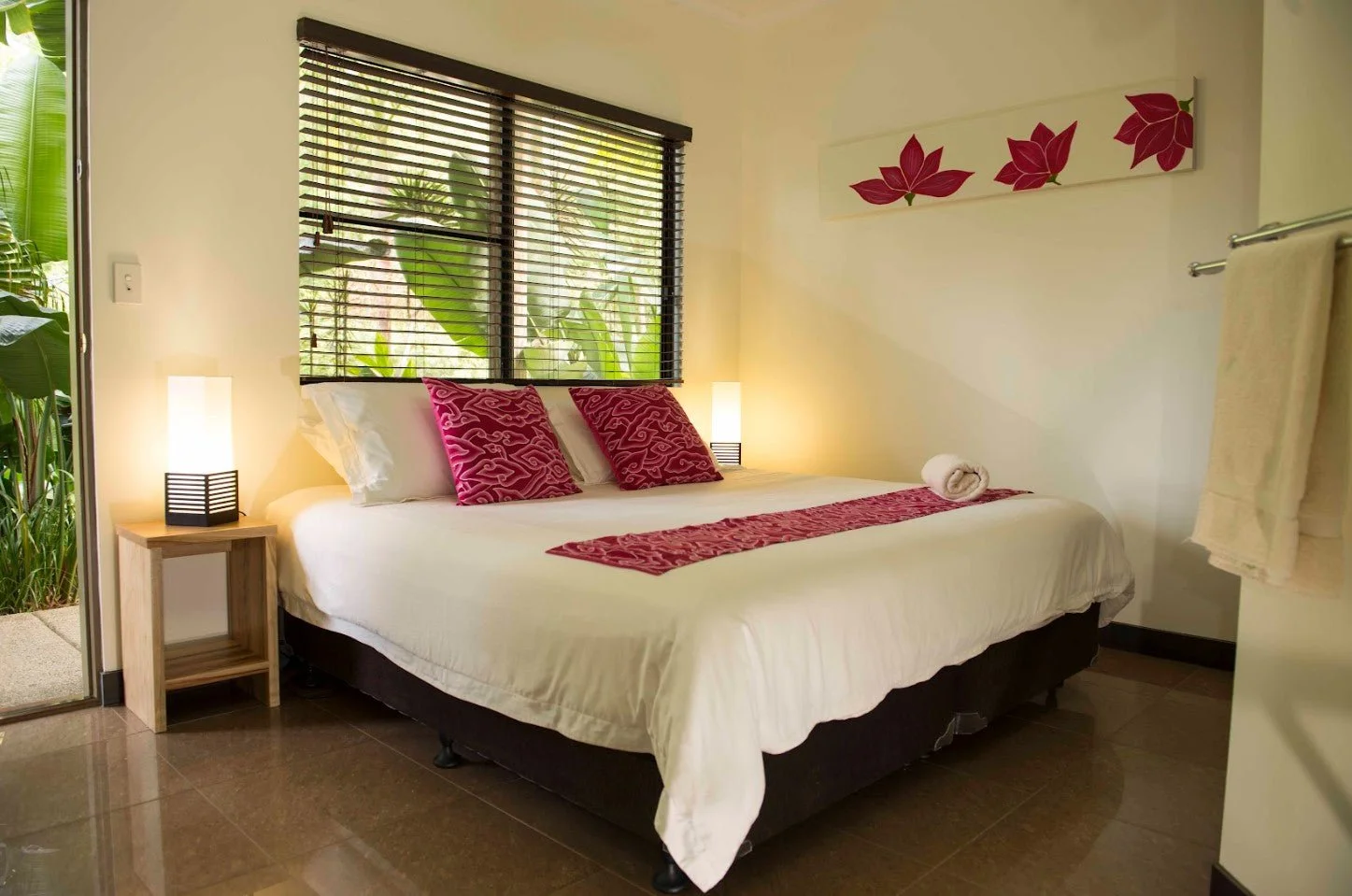 A bedroom with a large bed, pink pillows, and a white rolled towel. There is a window with blinds showing greenery outside, two bedside lamps emitting warm light, and a wall art with pink flowers. A towel hangs on a rack to the right.