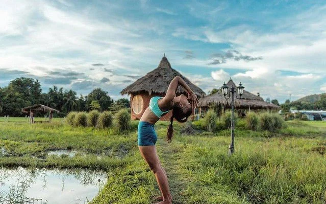 A woman practicing yoga outdoors near a pond and a traditional thatched-roof house on cloudy day.