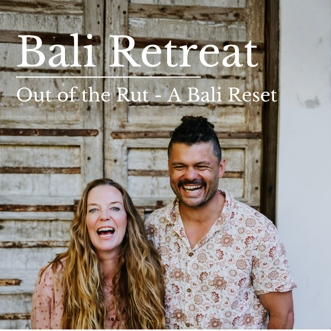 A smiling woman with long wavy hair and a smiling man with short dark hair and a patterned shirt standing in front of a rustic wooden wall during a Bali retreat.