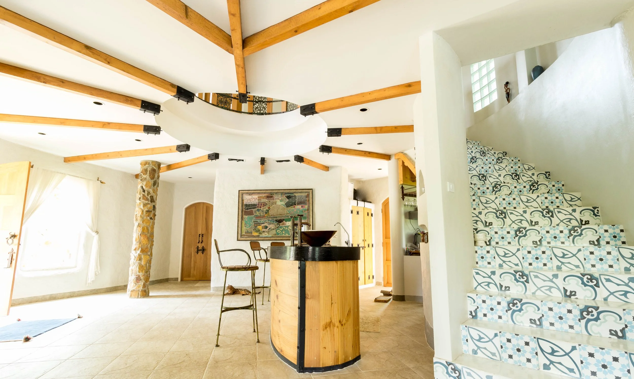 Interior view of a living space with a staircase with patterned tiles, a stone pillar, wooden beams on the ceiling, a yellow cabinet, a framed artwork, a kitchen island with a dark countertop, and a small dog resting on the floor.