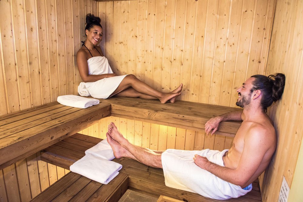 A man and woman sitting inside a wooden sauna, both wrapped in white towels, smiling and engaging in conversation.