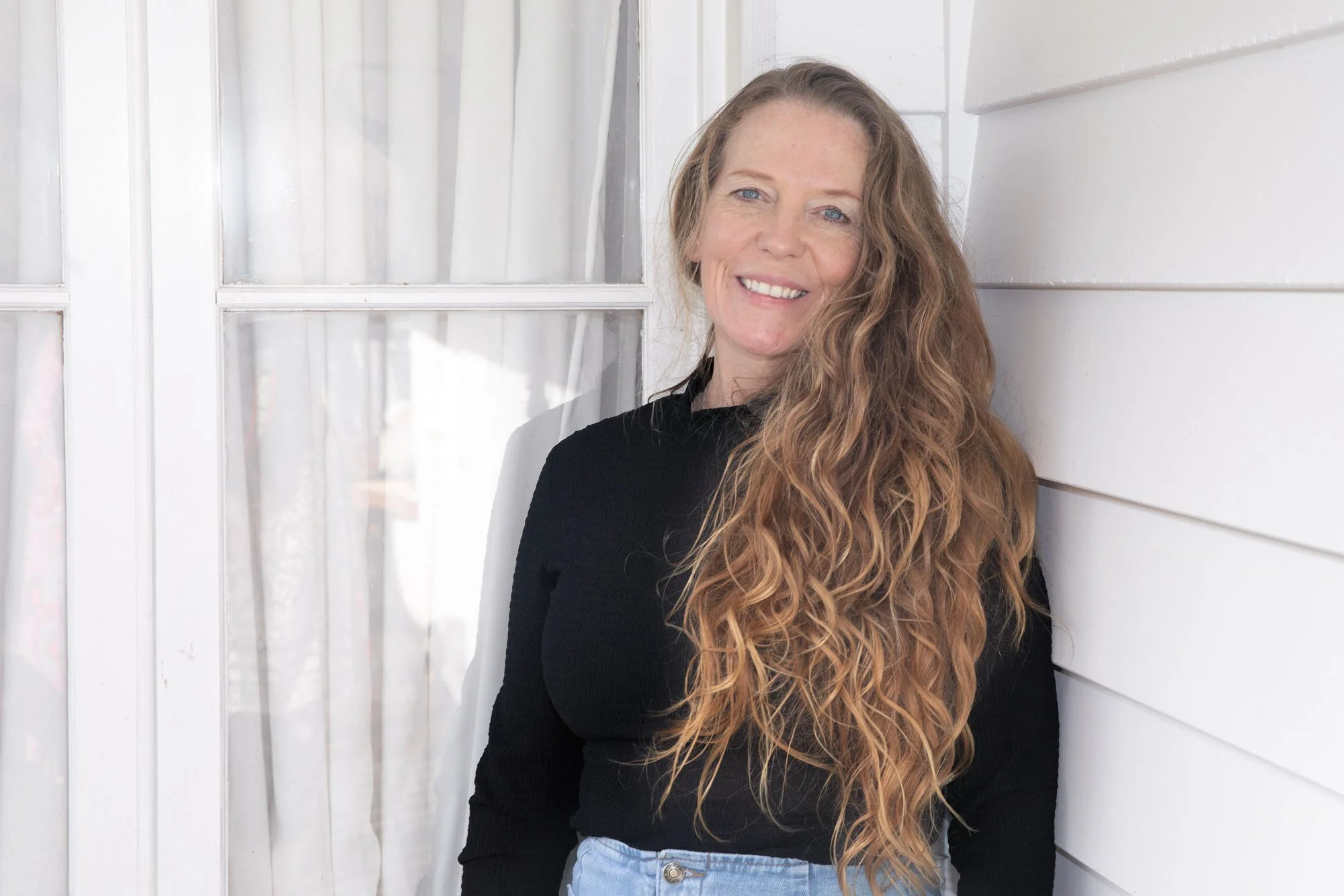 Woman with long, curly red hair, smiling, wearing a black top and light blue jeans, standing against a white wooden wall and window.