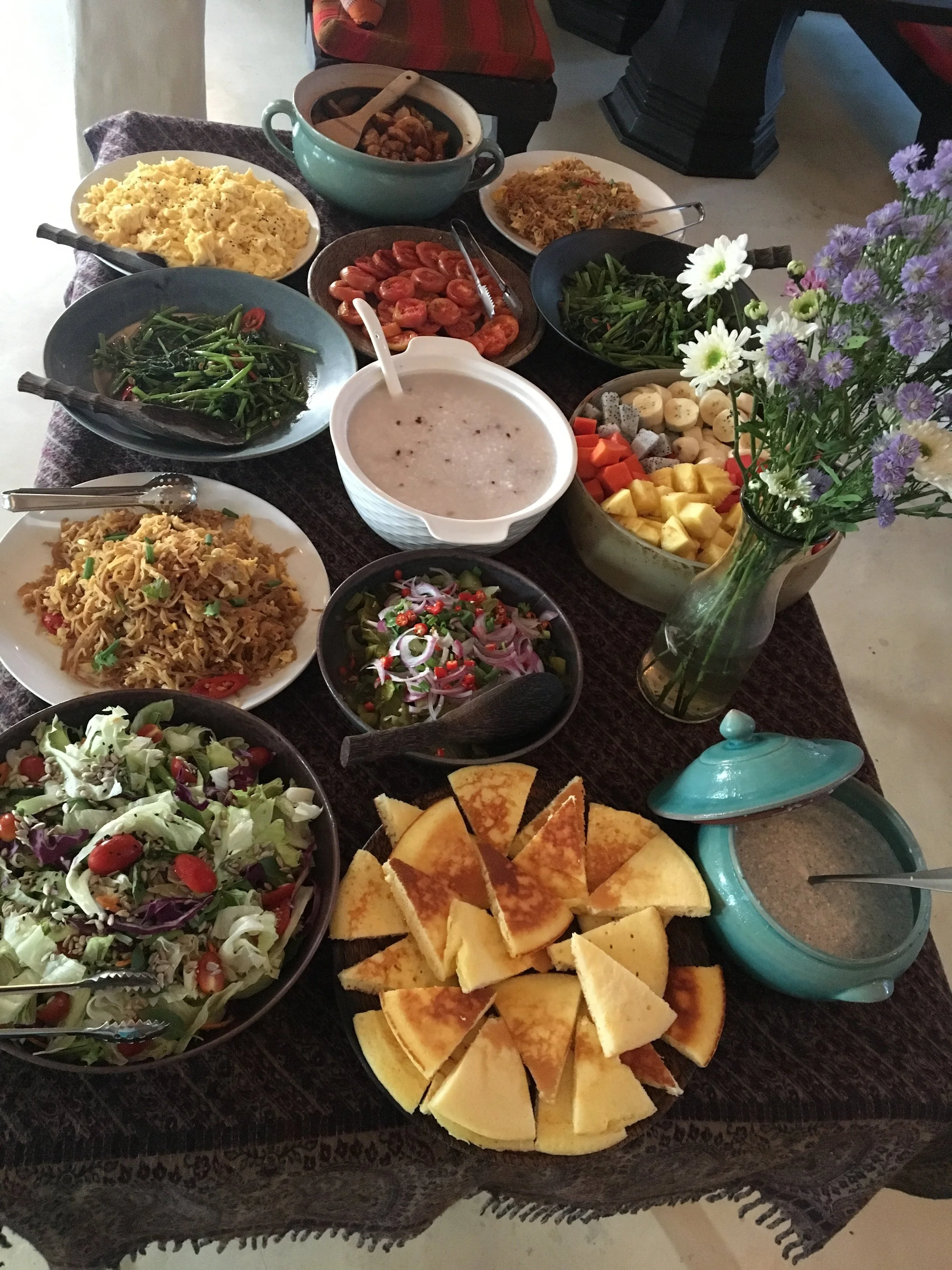 A variety of dishes and snacks on a dark table, including scrambled eggs, a bowl of rice, sliced tomatoes, a salad with cherry tomatoes and lettuce, a bowl of creamy soup with black pepper, a plate of sliced cake or bread, a bowl of noodles, a bowl o