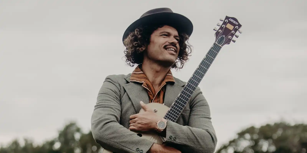 A man with curly hair, wearing a hat and a gray blazer, holding a guitar outdoors on a cloudy day.