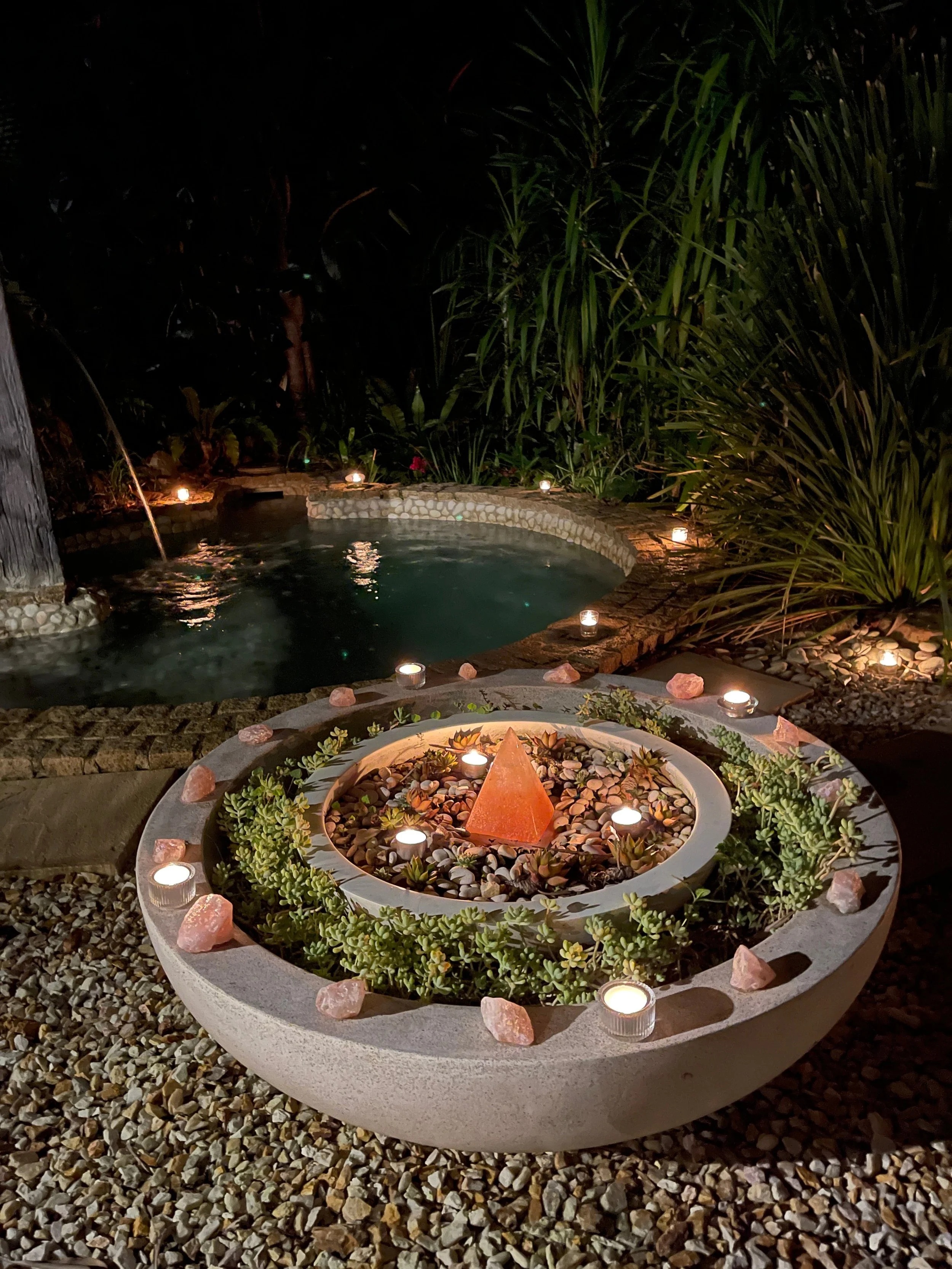 Nighttime outdoor scene with a small hot tub surrounded by candles, a decorative circular planter with rocks and succulents, and lush green foliage in the background.