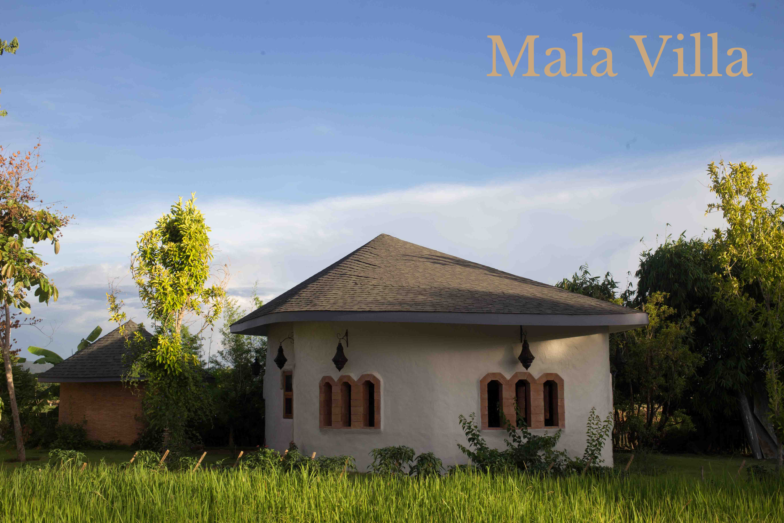 A small, white house with a rounded roof, surrounded by green grass and trees, under a clear blue sky with the text "Mala Villa" in the top right corner.