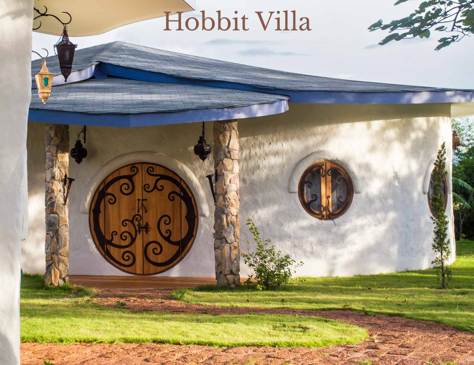 A whimsical house with a curved roof and white stucco walls, featuring a large round wooden door with black wrought-iron swirl patterns, and two smaller round windows with matching wrought-iron grilles, surrounded by a green lawn and small trees.