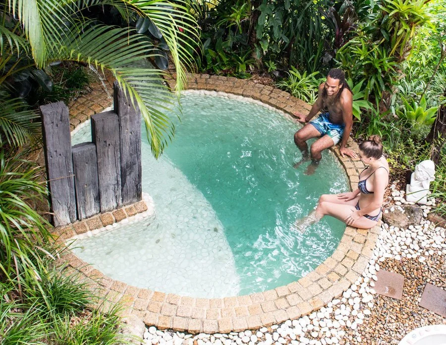 Two people relaxing in a small, round backyard hot tub surrounded by lush tropical plants, one woman in a bikini sitting on the edge and a man sitting inside the hot tub.