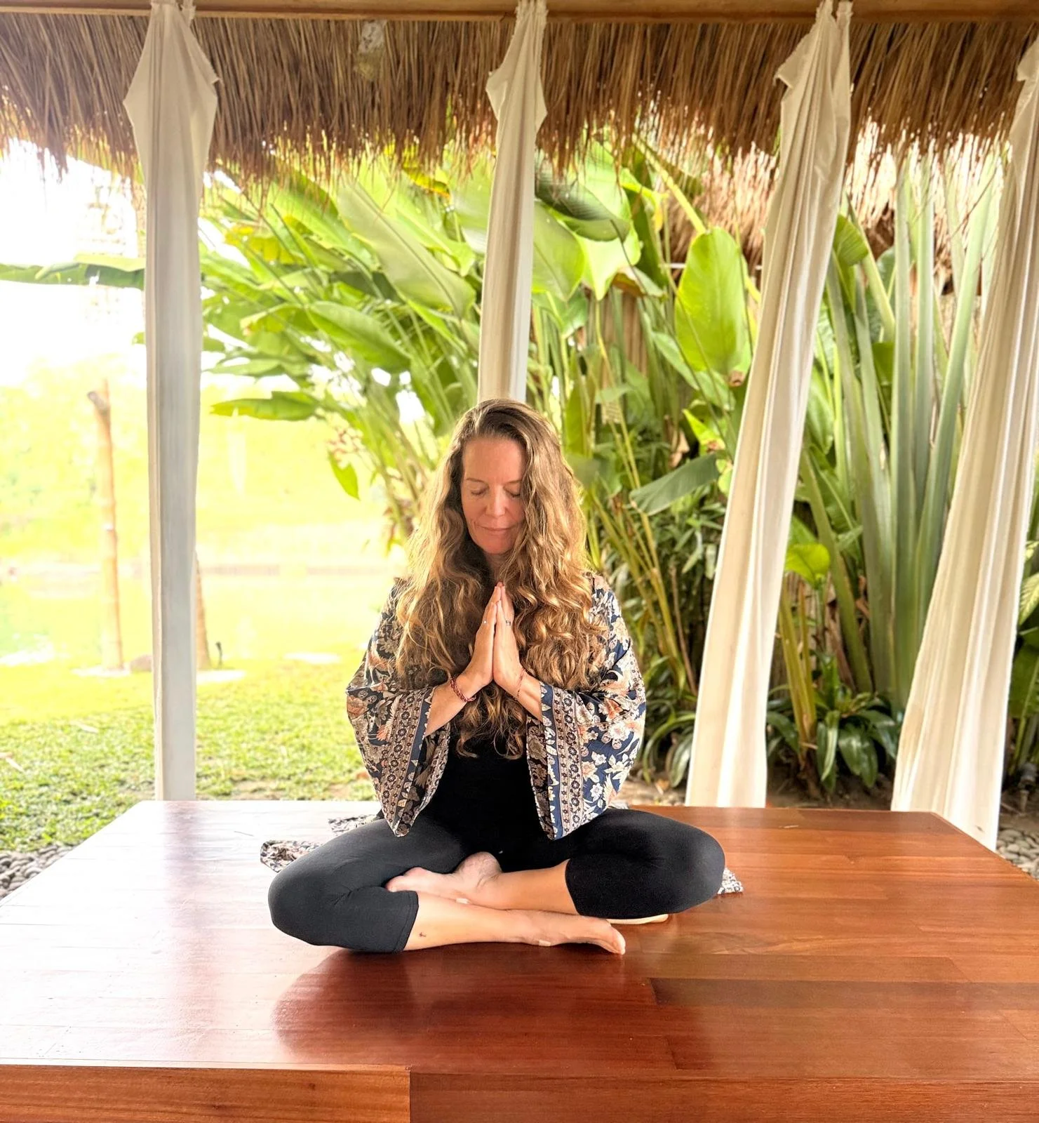 A woman practicing yoga or meditation outdoors, seated cross-legged on a wooden platform, with her hands in prayer position, surrounded by green plants and a natural setting.