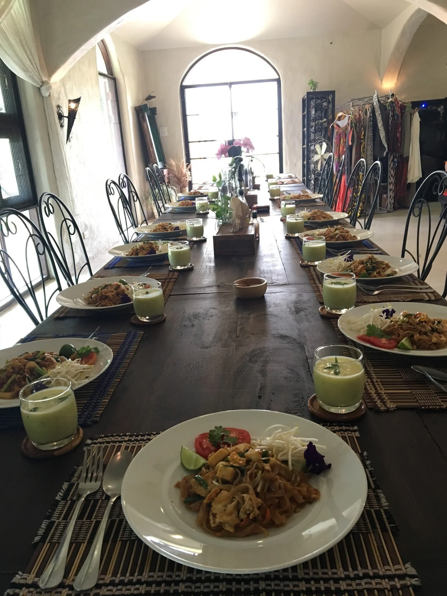 A long dining table set for a meal with plates of noodles, vegetables, lime wedges, and green drinks, in a room with arched windows and decorative clothing and plants in the background.
