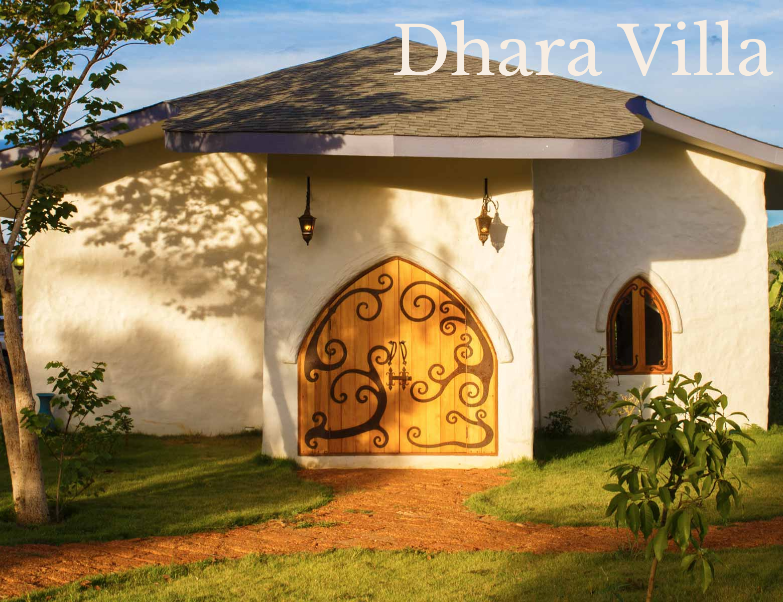 White stucco building with a rounded wooden door featuring black scrollwork, a small arched window, and outdoor lanterns, surrounded by greenery.