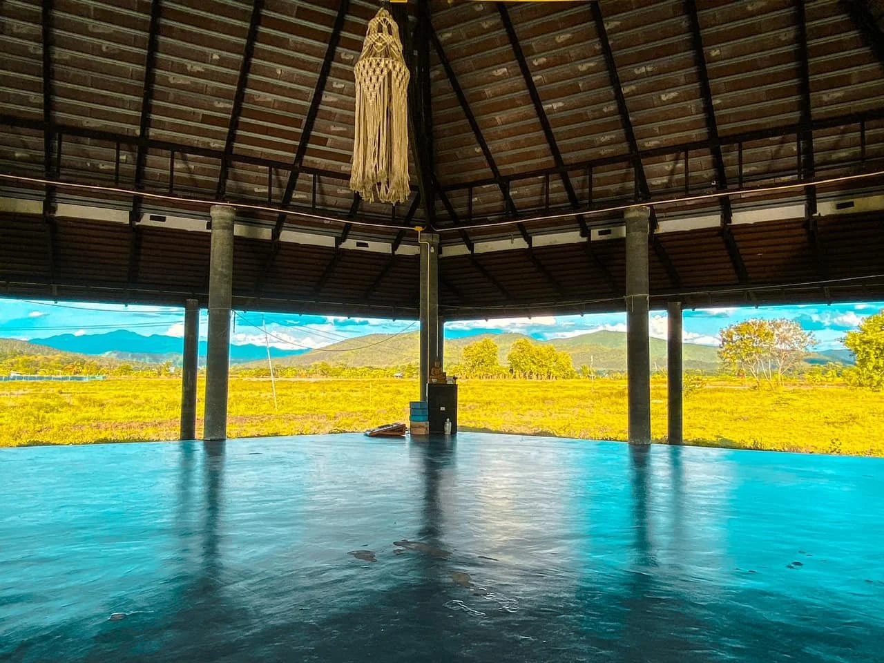 Open-air pavilion with a thatched roof, overlooking a scenic landscape of fields, trees, and mountains under a blue sky.