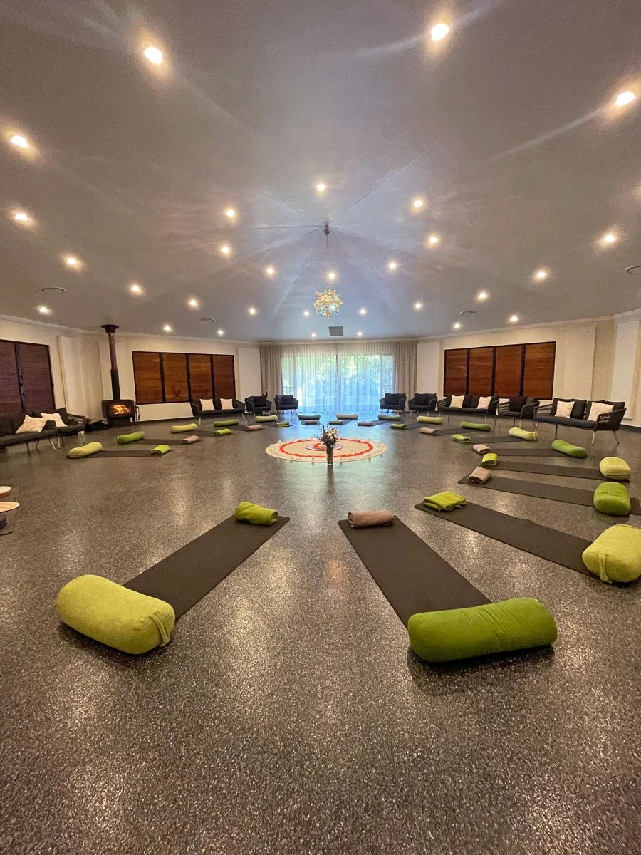 A spacious room set up for a yoga or meditation class with black mats, green bolsters, and small pillows arranged in a circle. The room has a polished dark floor, large windows with sheer curtains, and warm overhead lighting. There is a small decorat