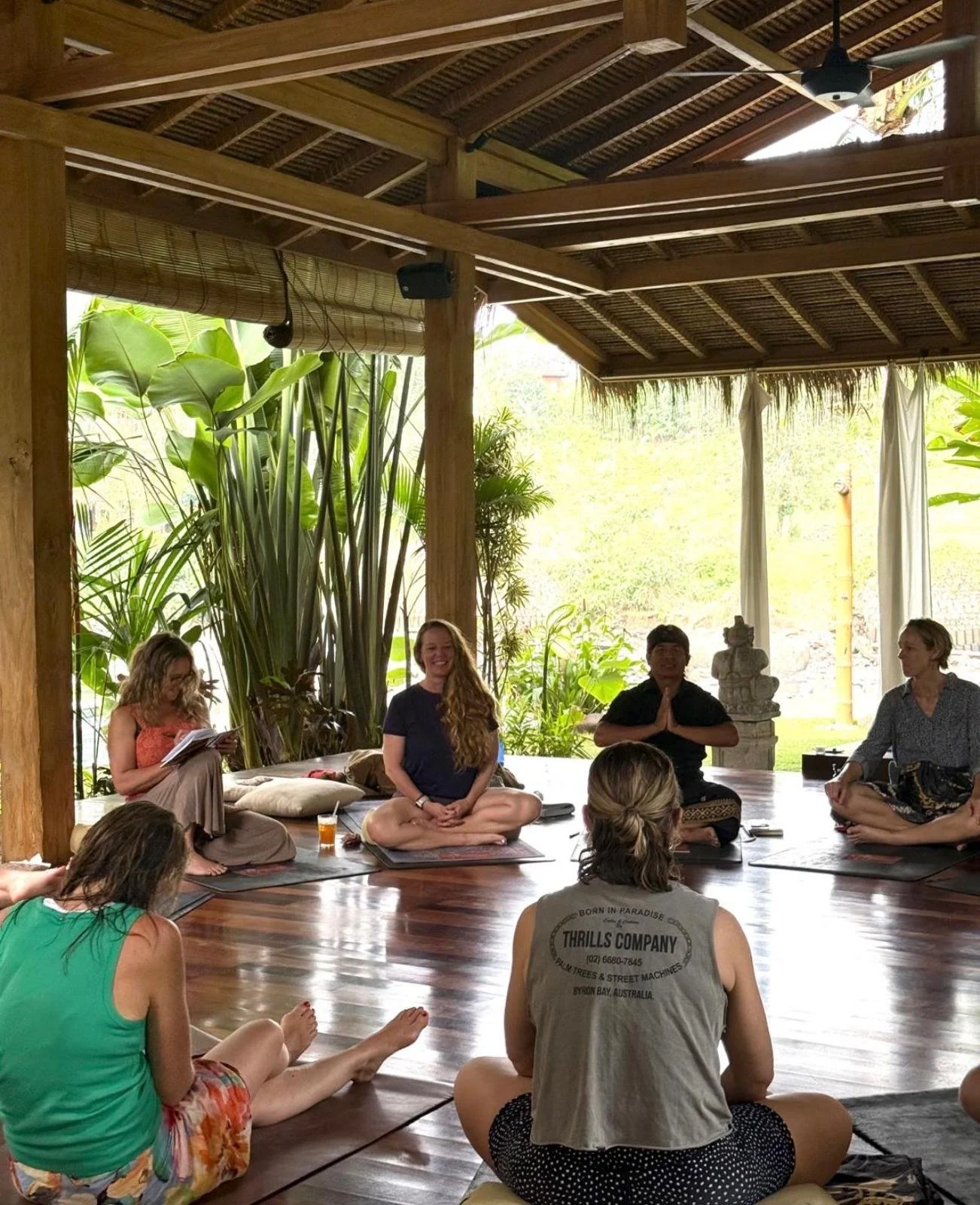 People participating in a meditation or yoga session in a tropical open-air setting with lush greenery and wooden structure.