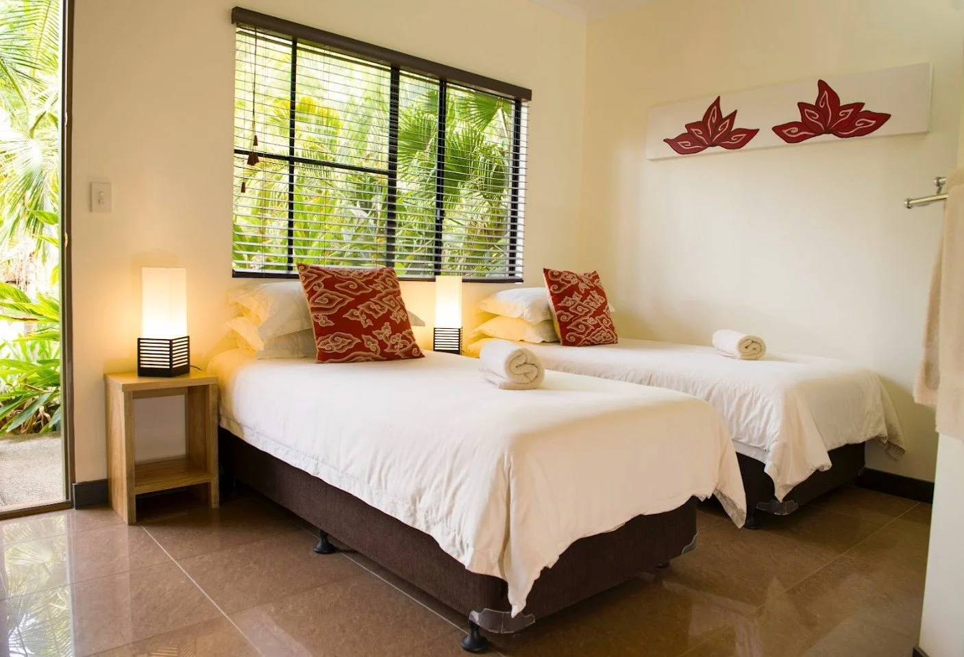 A bedroom with two beds, each with red patterned pillows and rolled white towels at the foot. There are two bedside lamps on wooden tables. A window with black blinds shows green foliage outside. A white wall with red decorative artwork is above the 